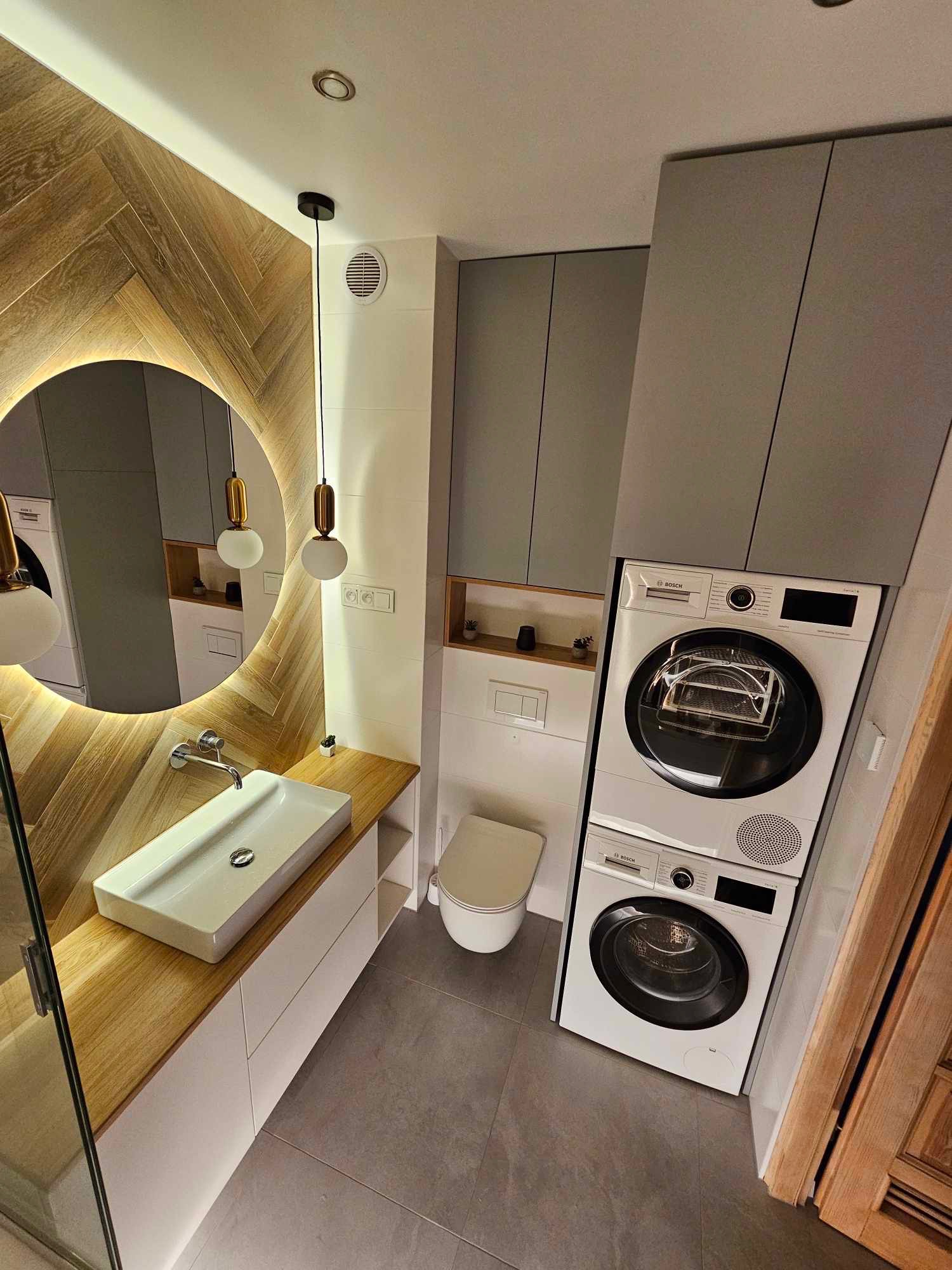 Nowoczesna łazienka z okrągłym lustrem podświetlanym LED, drewnianym blatem i szafkami w odcieniach bieli i szarości. Uwagę przyciąga ściana wykończona drewnem ułożonym w jodełkę.