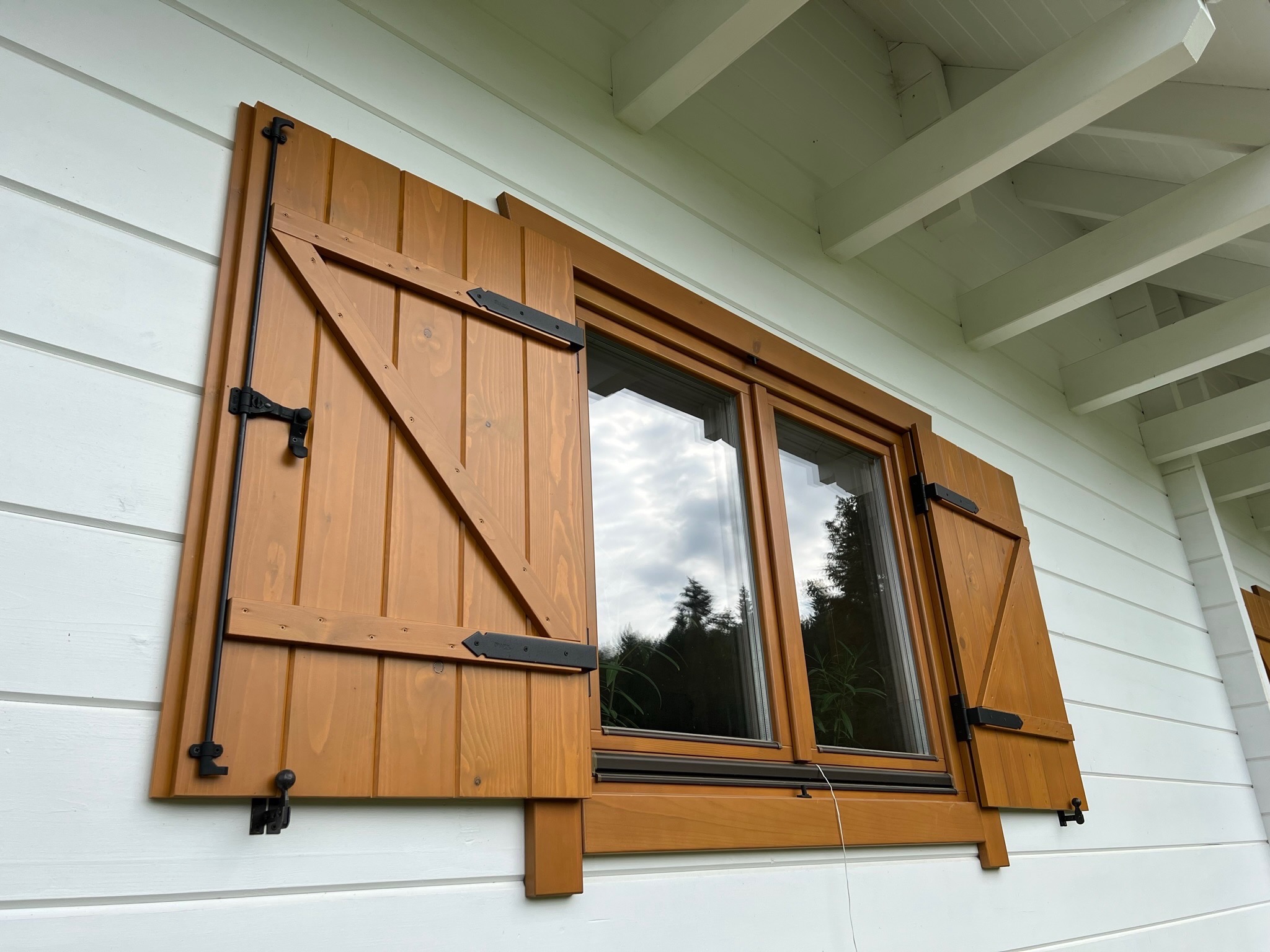 Drewniane okno z otwartymi okiennicami w rustykalnym stylu, widoczne czarne okucia. Biała elewacja domu z drewnianą podbitką dachową.