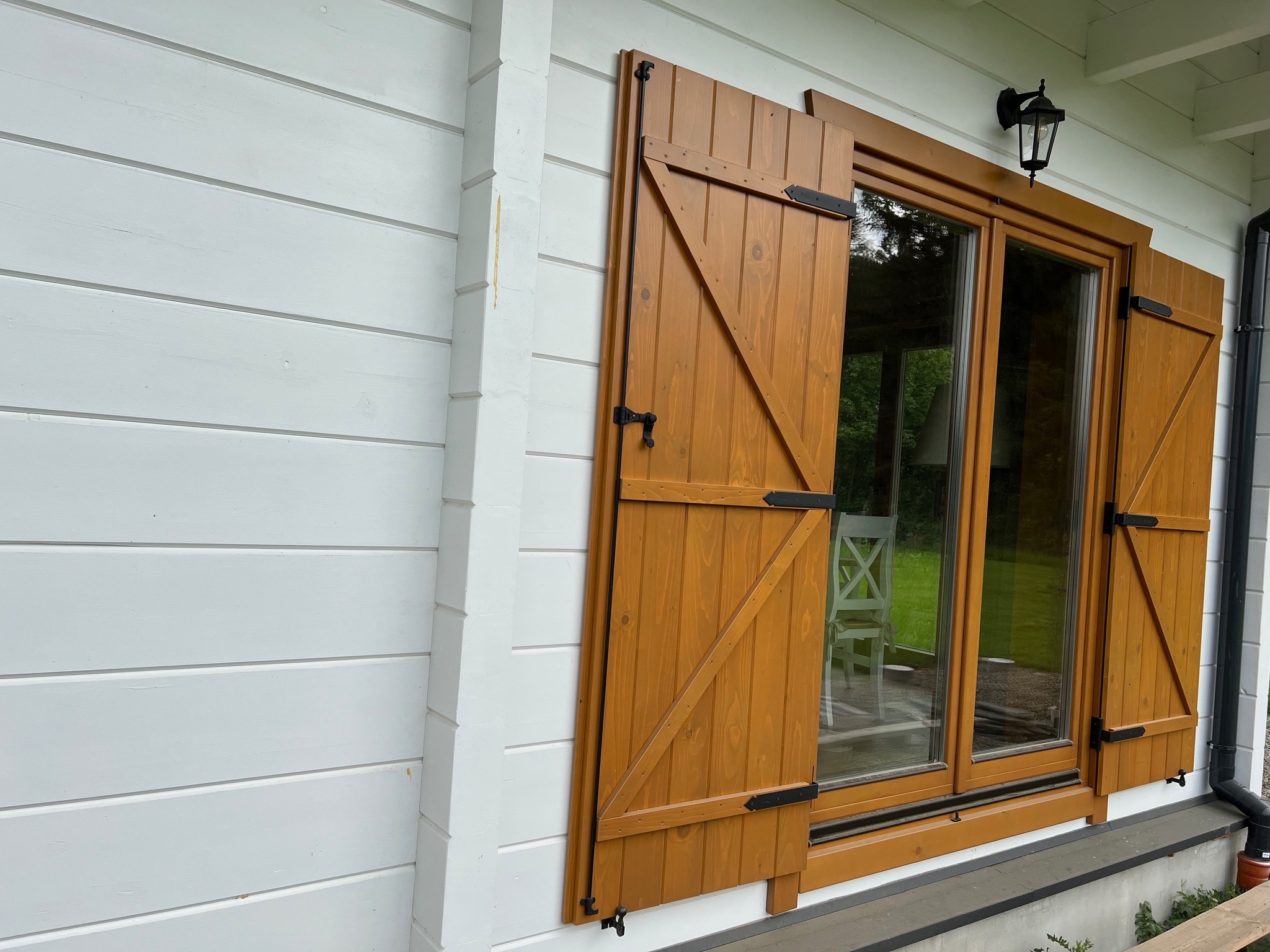 Drewniane okiennice w kolorze miodowym z czarnymi okuciami, zamontowane na białej, drewnianej elewacji domu. Widoczne okno z ramą w tym samym kolorze co okiennice.