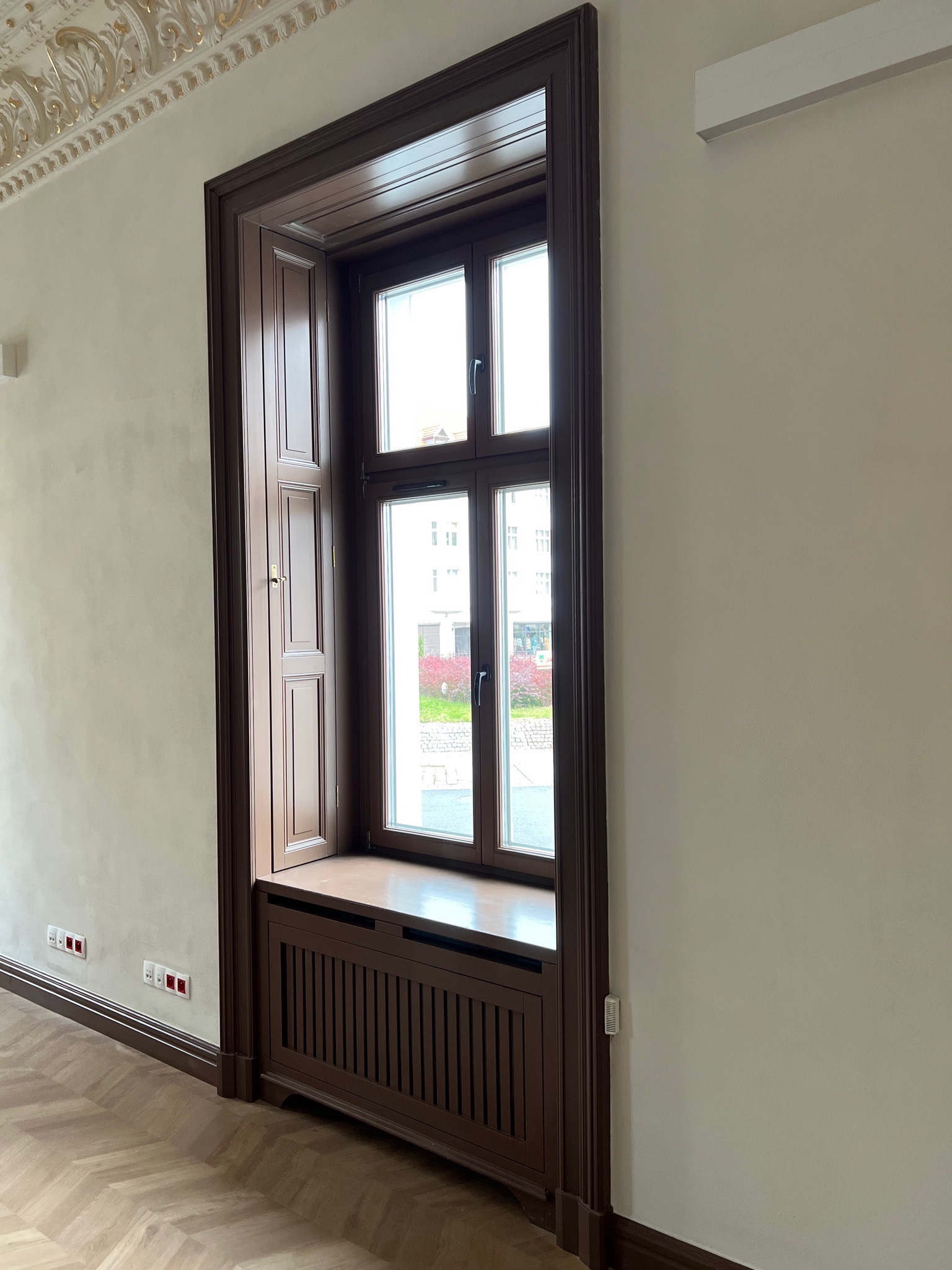 Stylowe okno drewniane z okiennicami w ciemnej ramie, wkomponowane w ścianę z dekoracyjnym sufitem i parkietem w jodełkę. Widoczny grzejnik pod oknem.