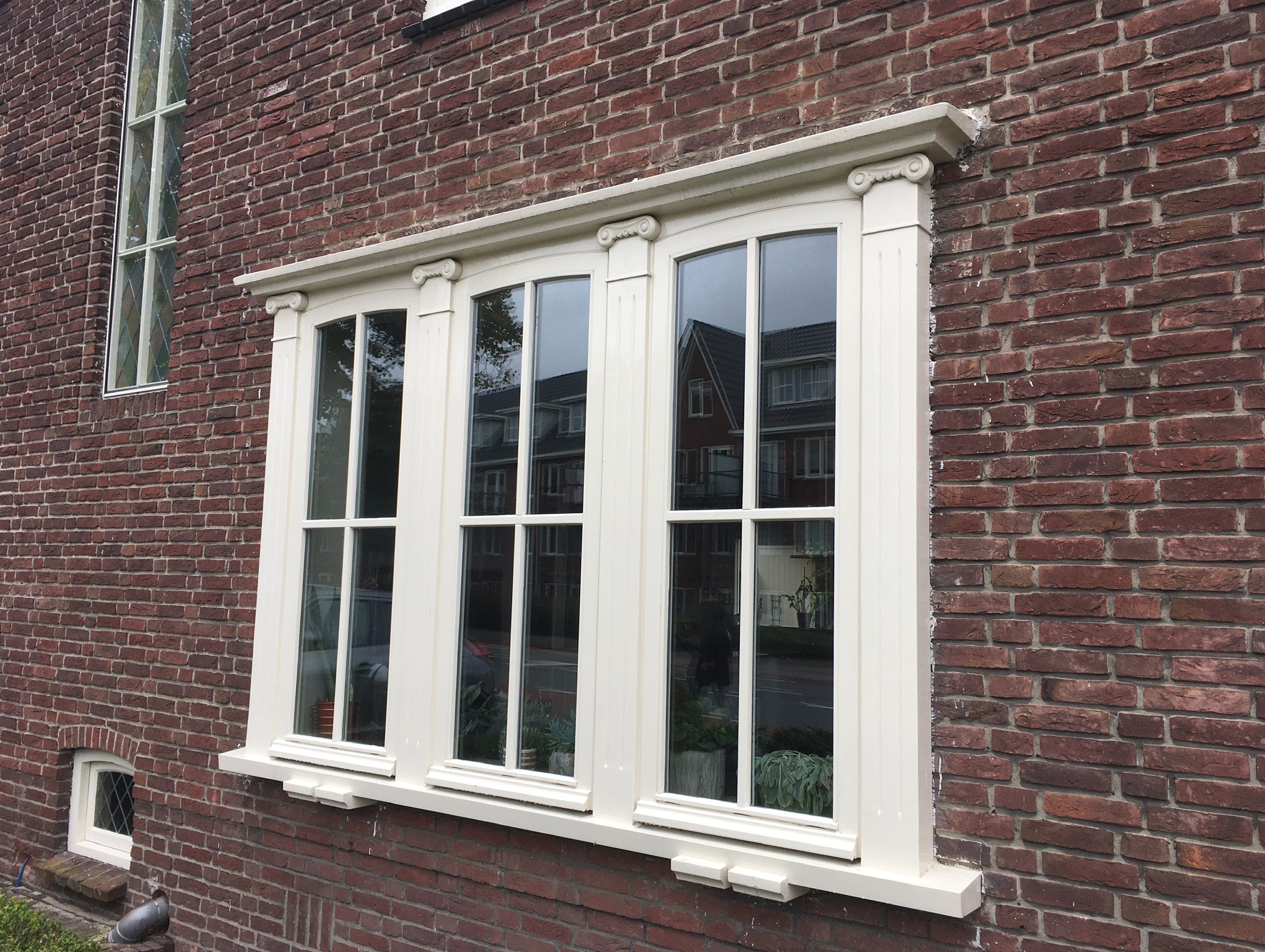 Eleganckie, białe okno drewniane z trzema skrzydłami, zdobione detalami architektonicznymi, wkomponowane w czerwoną ceglaną ścianę budynku. Widoczne odbicie otoczenia w szybach.