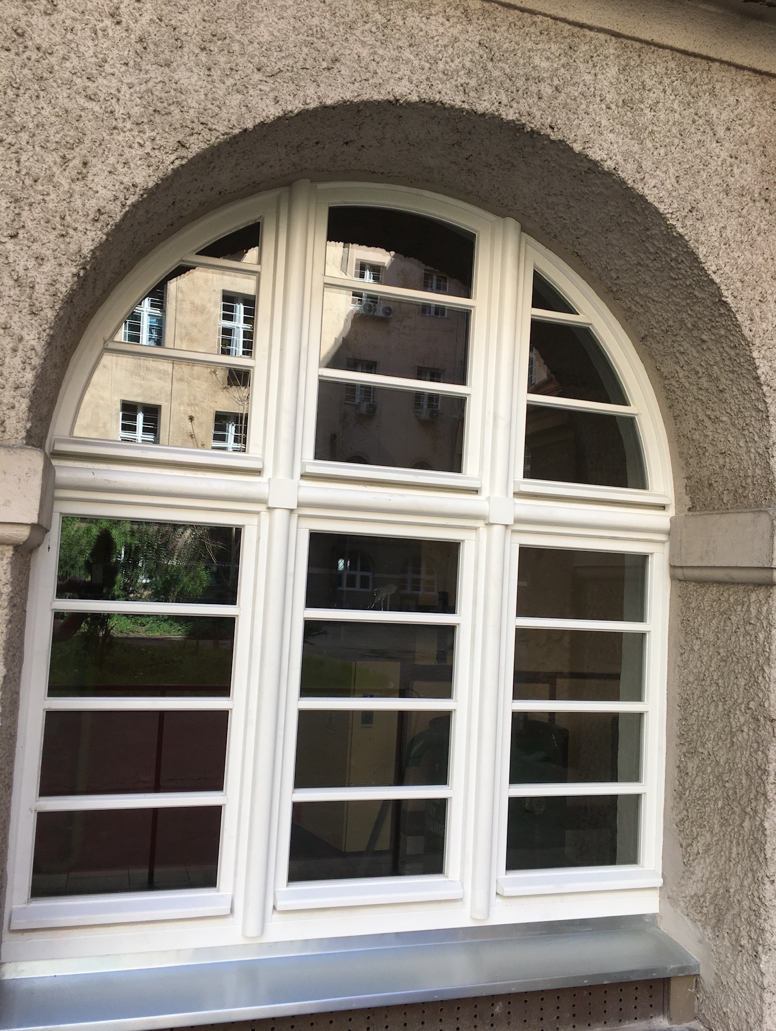 Białe, łukowe okno drewniane z podziałami, osadzone w szarej, teksturowanej ścianie. W szybach odbija się budynek i zieleń.
