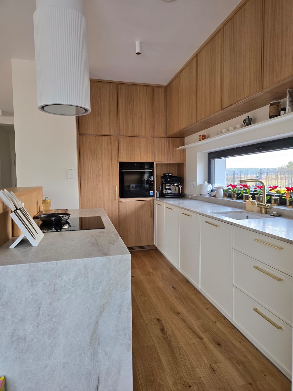 Nowoczesna kuchnia z drewnianymi frontami szafek górnych i białymi dolnymi, złotymi uchwytami, marmurowym blatem i drewnianą podłogą. Widoczny okap cylindryczny.