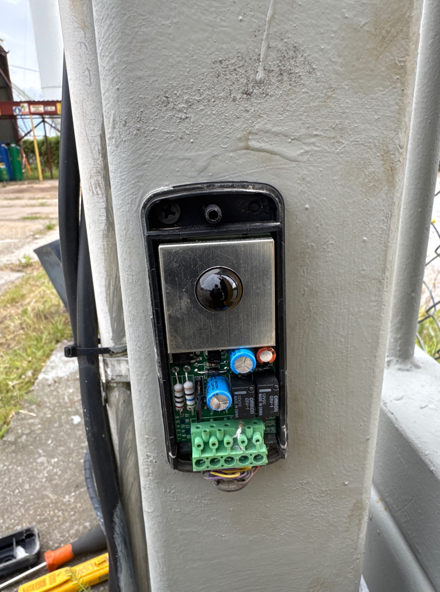 Otwarta obudowa czujnika z widoczną elektroniką, zamontowana na szarym słupku. Widoczne przewody, płytka drukowana z elementami i złącza.