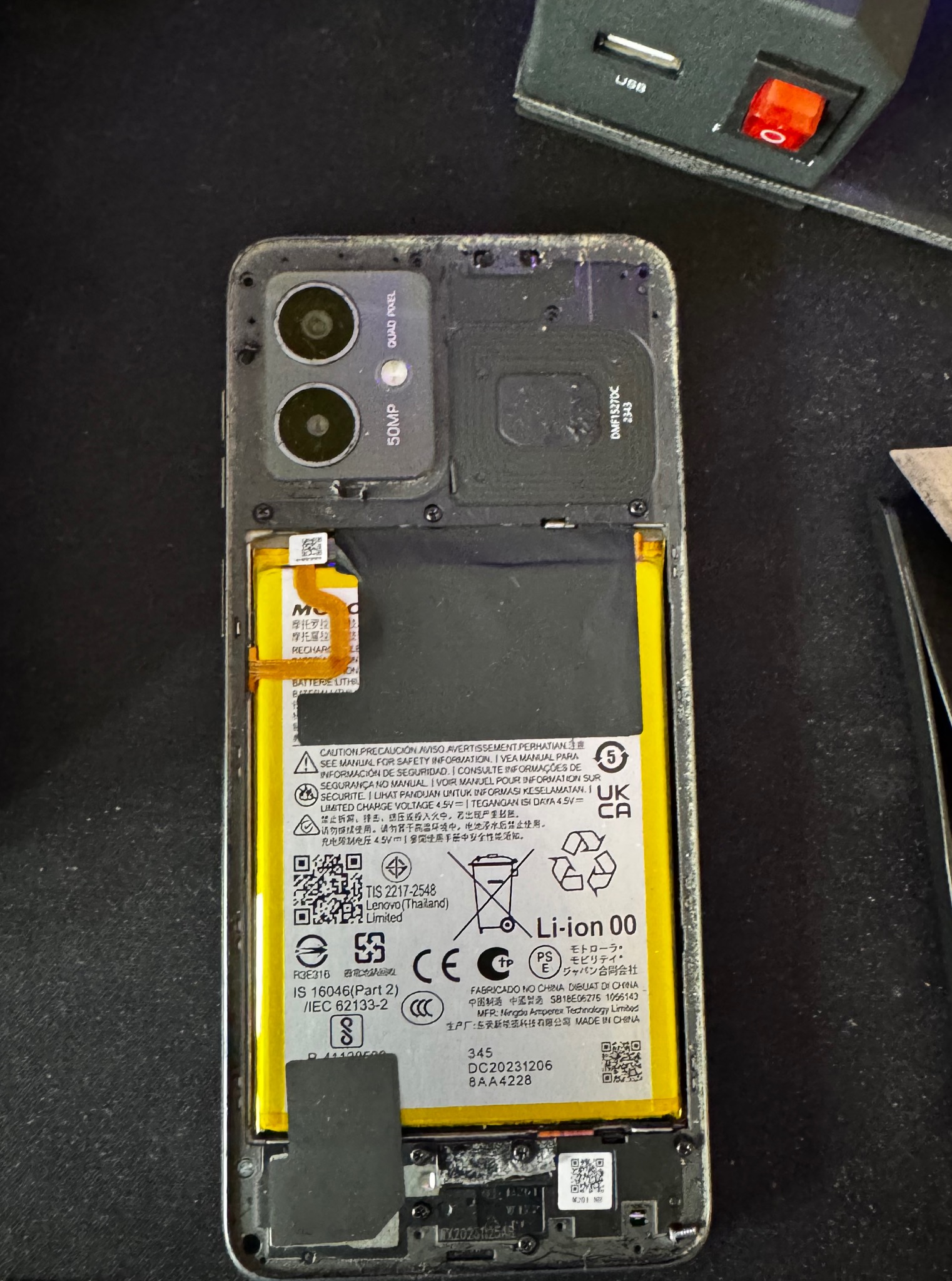 Otwarty smartfon z widoczną baterią i wewnętrznymi komponentami. Widoczne napisy i oznaczenia na baterii. W tle narzędzia serwisowe.