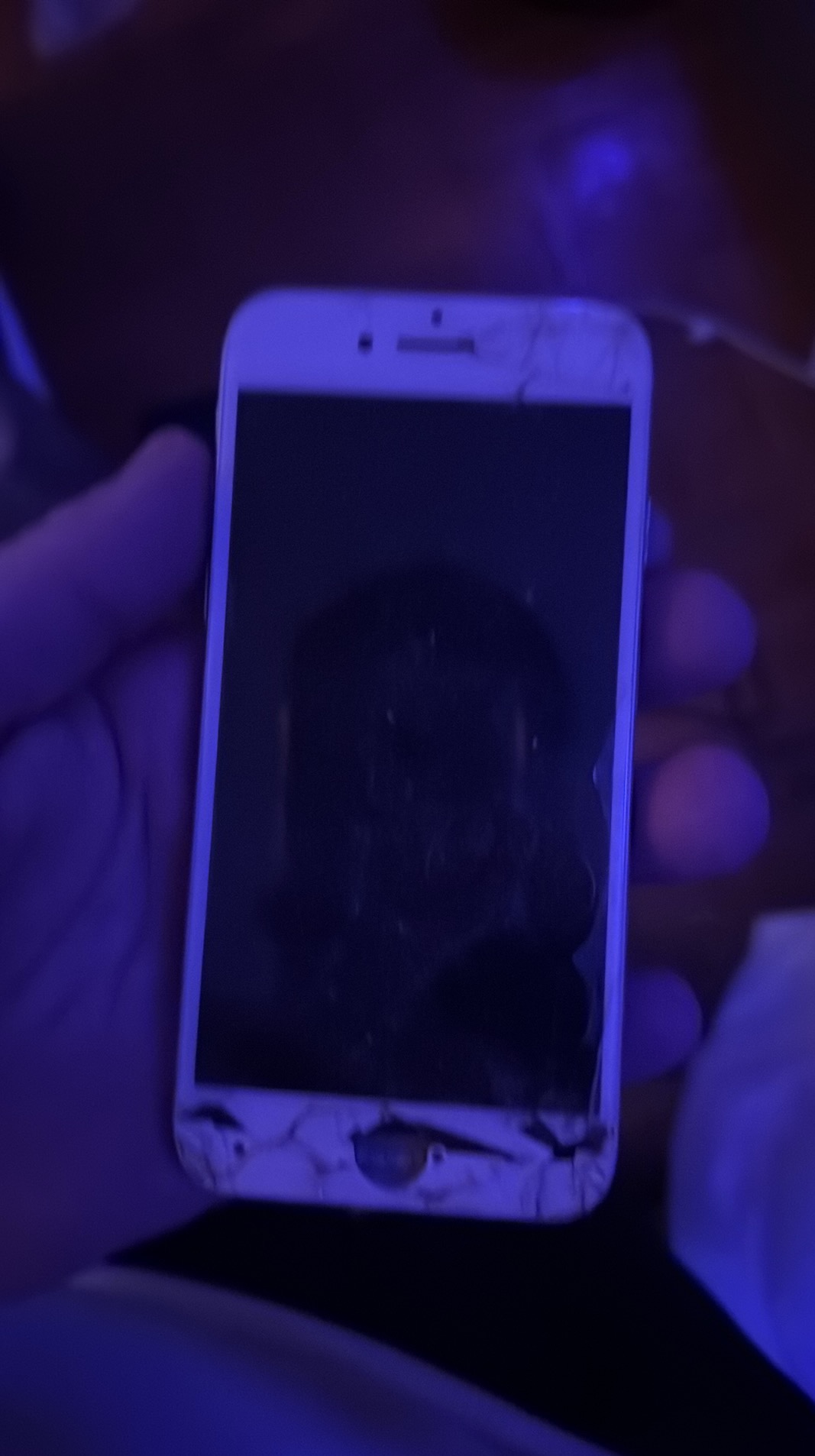 Dłoń trzyma uszkodzony, biały smartfon z pękniętym ekranem i obudową. Widoczne ślady użytkowania i uszkodzenia mechaniczne. Ujęcie w słabym, niebieskawym oświetleniu.