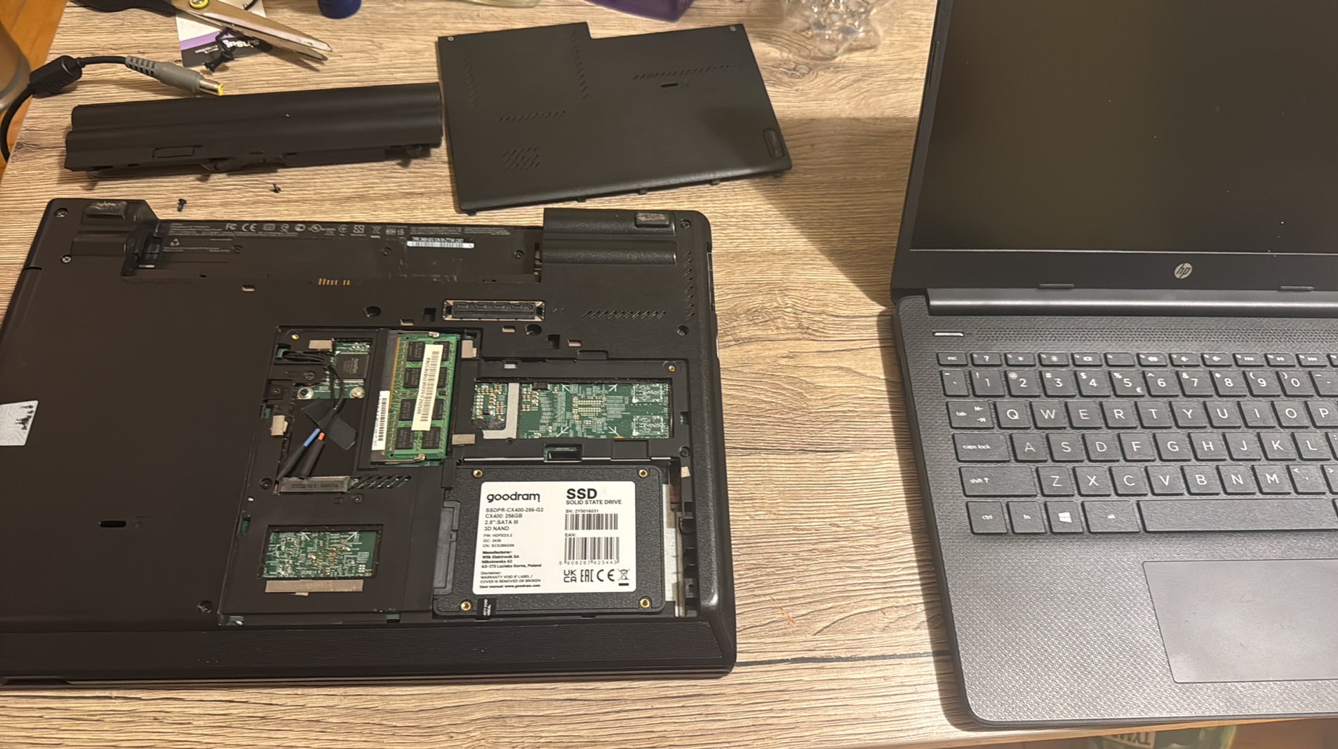Otwarty laptop z widocznymi podzespołami, dyskiem SSD Goodram, obok drugi laptop HP. Widoczne narzędzia i bateria. Serwis laptopów, wymiana dysku.
