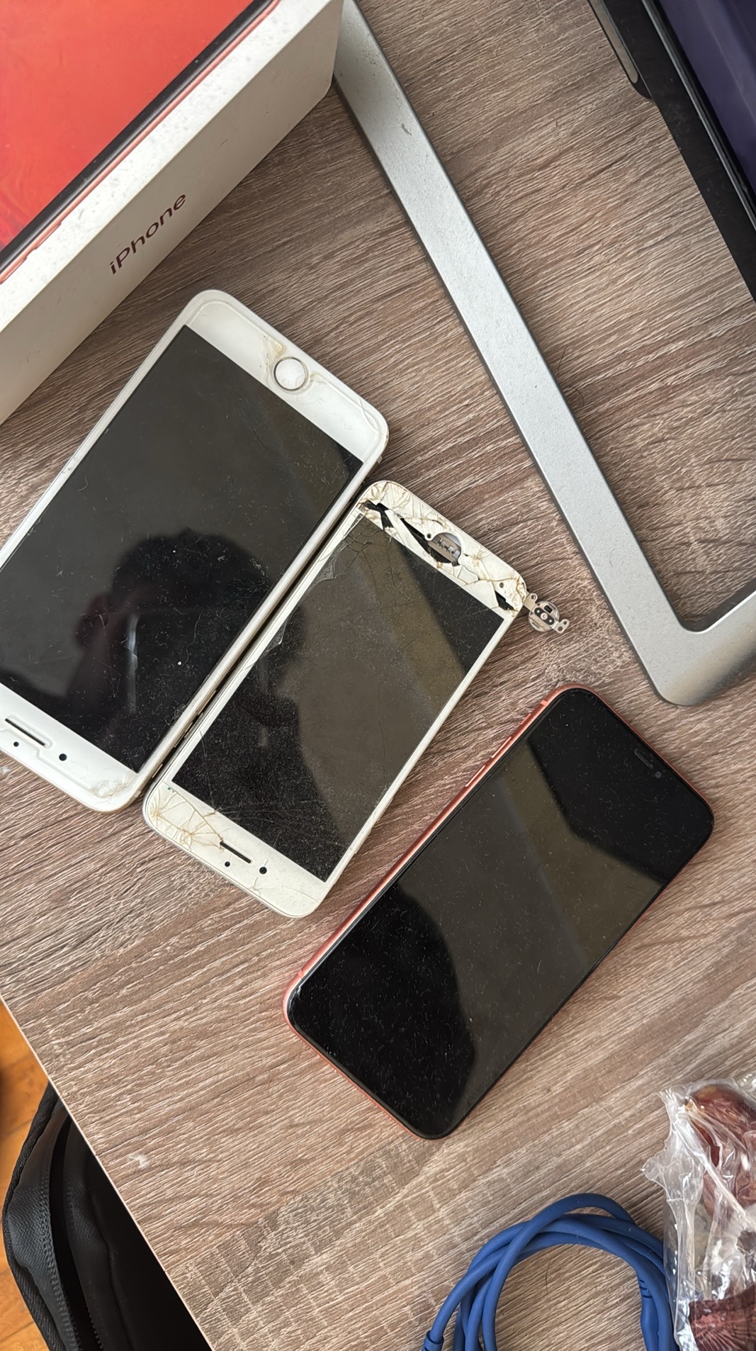 Uszkodzone telefony iPhone z pękniętymi ekranami leżące na stole, obok pudełko i narzędzia. Widoczne ślady użytkowania i uszkodzenia mechaniczne.