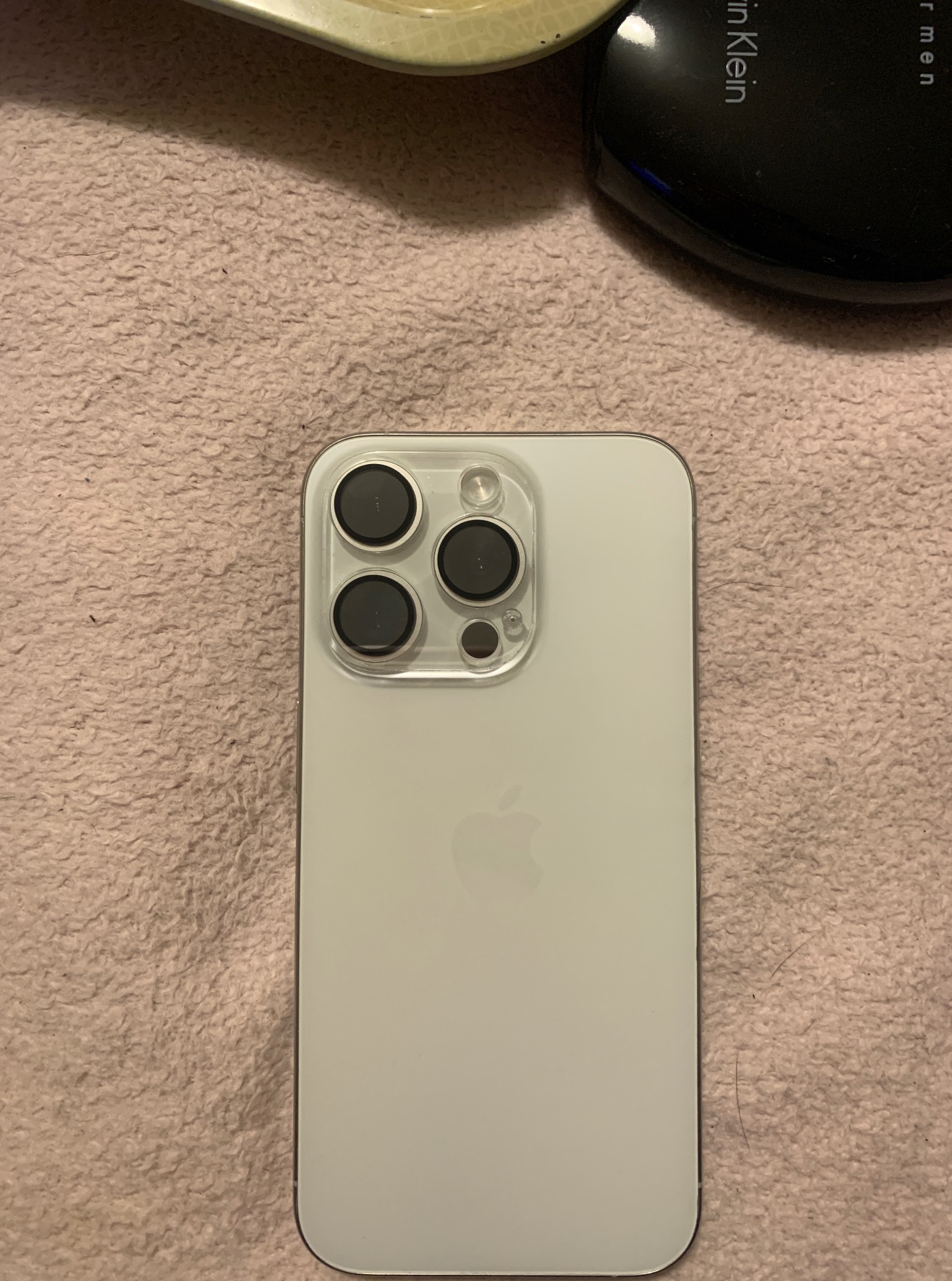 Biały iPhone z trzema obiektywami na teksturowanym tle, obok czarnego pudełka kosmetycznego i beżowego przedmiotu. Widok z tyłu, skupienie na obiektywach.