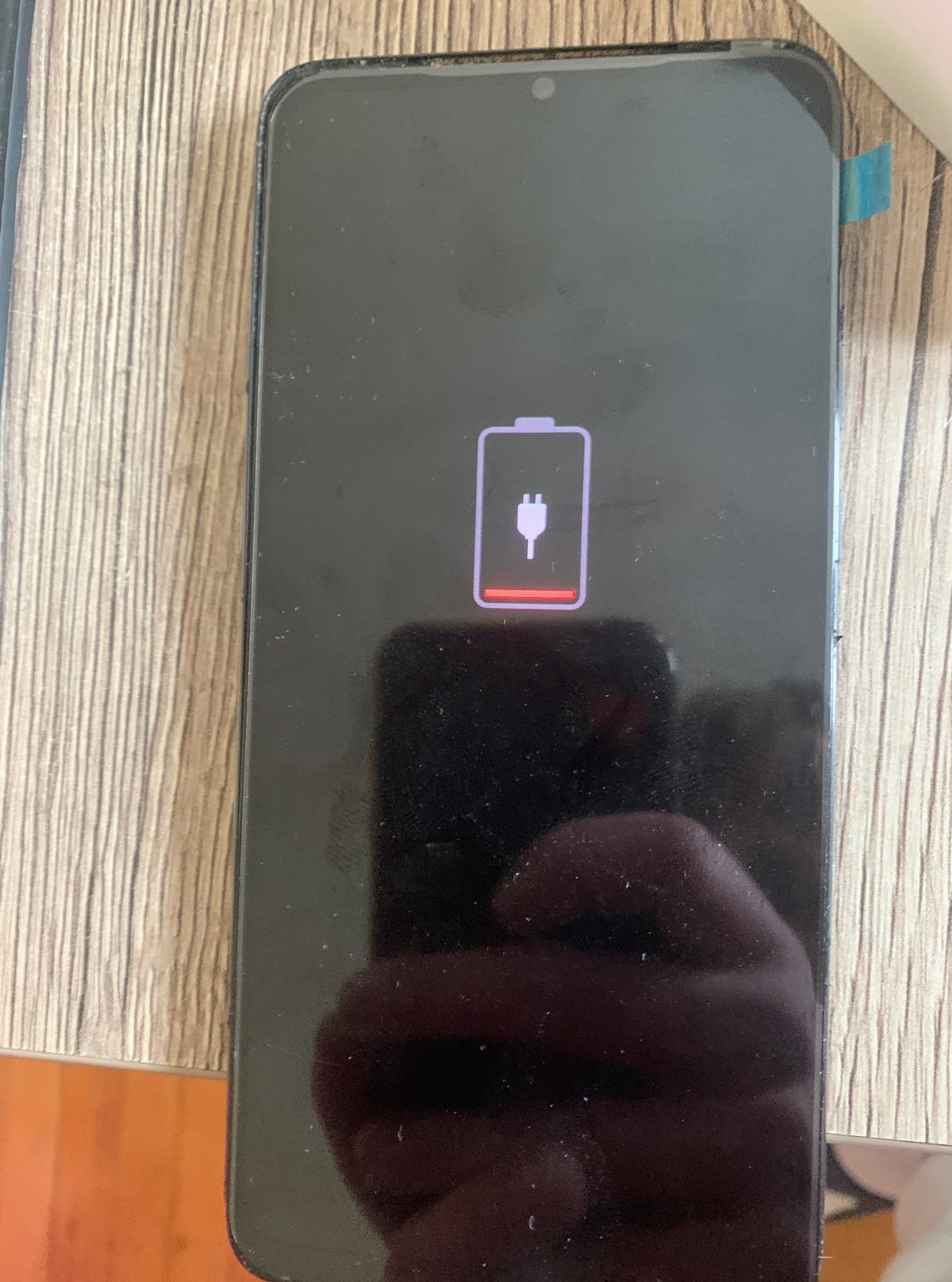 Czarny smartfon z ikoną rozładowanej baterii na ekranie, leżący na drewnianej powierzchni. Widoczny fragment niebieskiej taśmy na obudowie.