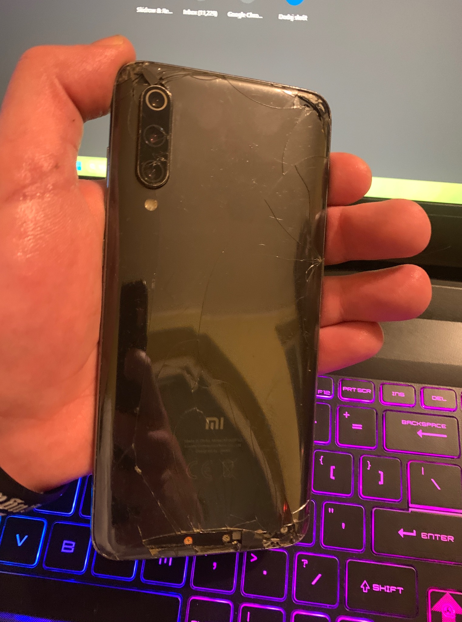 Czarny smartfon Xiaomi z pękniętą obudową, trzymany w dłoni nad podświetlaną klawiaturą laptopa. Widoczne uszkodzenia na tylnej obudowie i w okolicy kamery.
