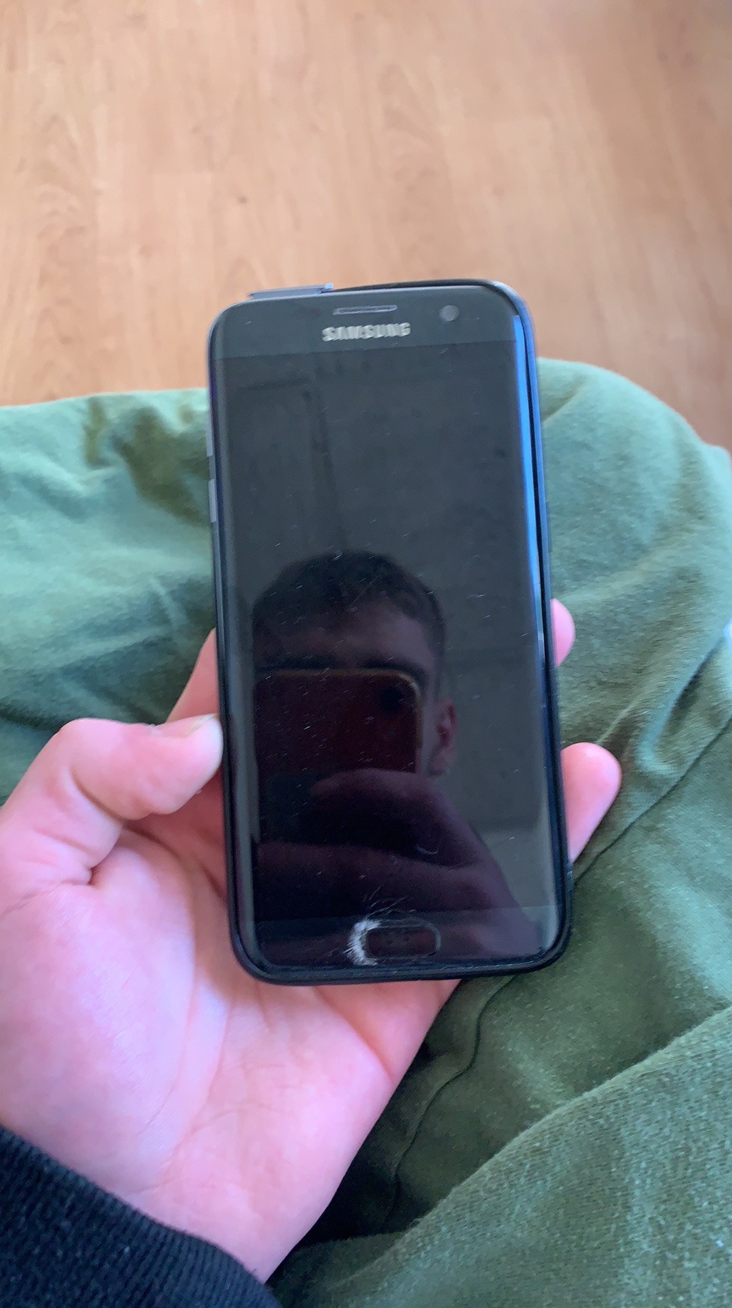 Czarny smartfon Samsung z pękniętym ekranem, trzymany w dłoni na tle zielonej tkaniny. Widoczne logo Samsung. Refleks w ekranie.