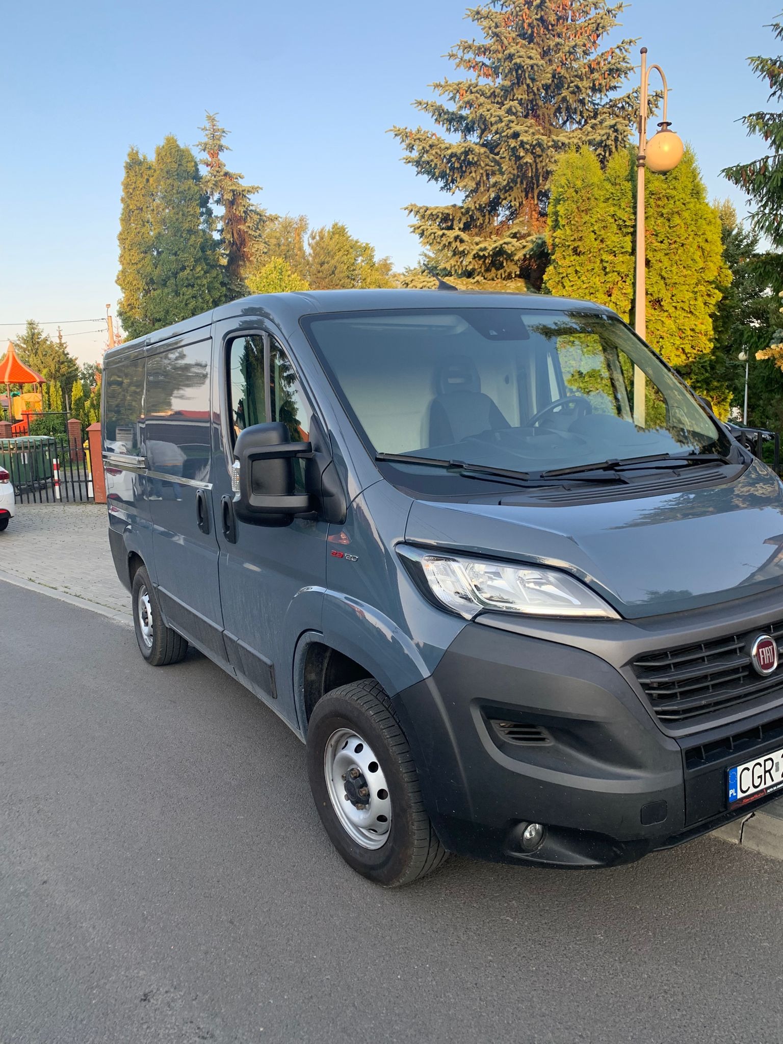 Szary bus Fiat Ducato zaparkowany na ulicy, z widocznym przodem i bokiem, na tle zieleni i placu zabaw. Tablica rejestracyjna widoczna.