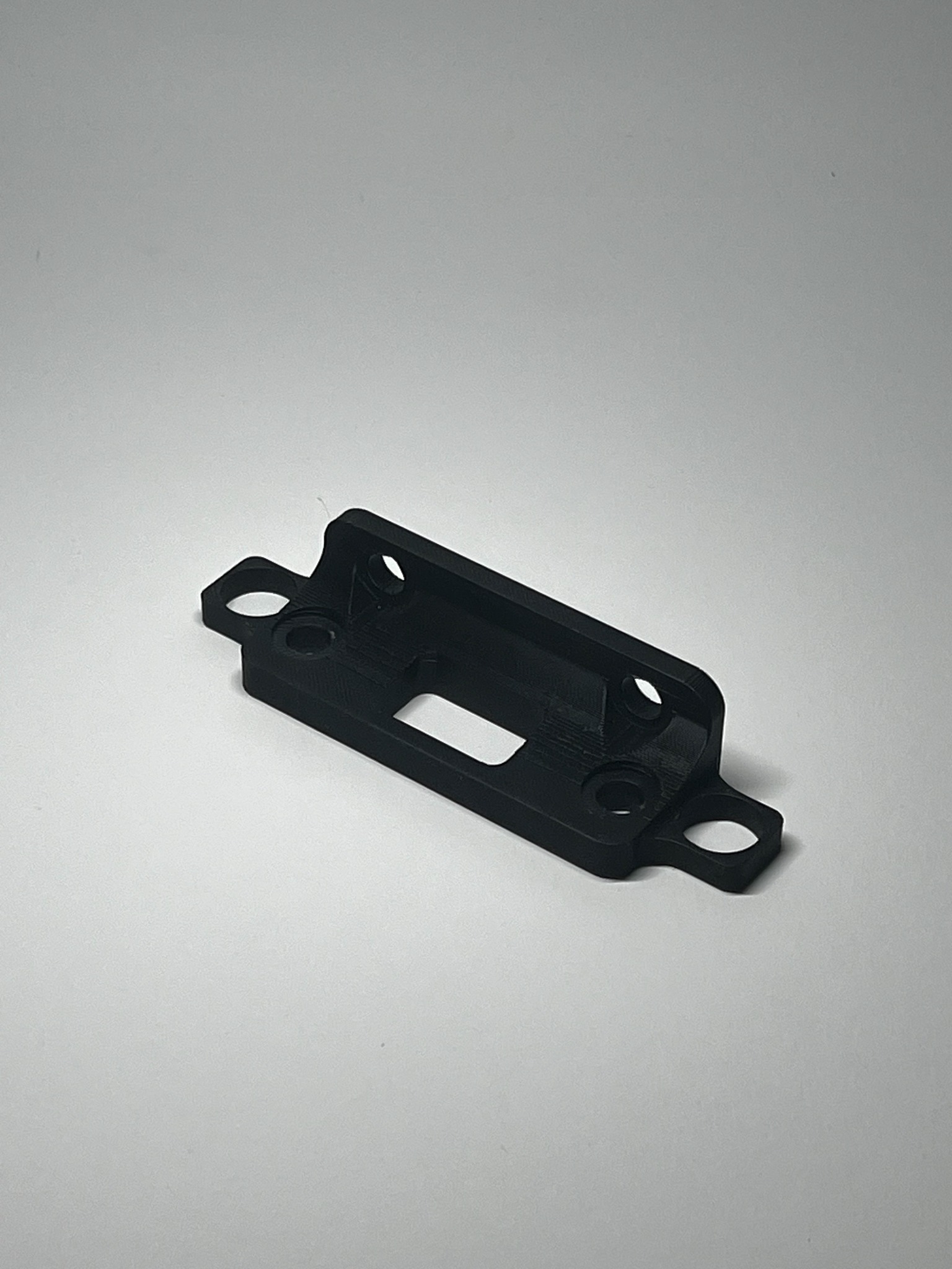 Czarny, matowy element z tworzywa sztucznego, wydrukowany w 3D, z otworami i prostokątnym wycięciem, leżący na białym tle. Widoczne delikatne warstwy druku.