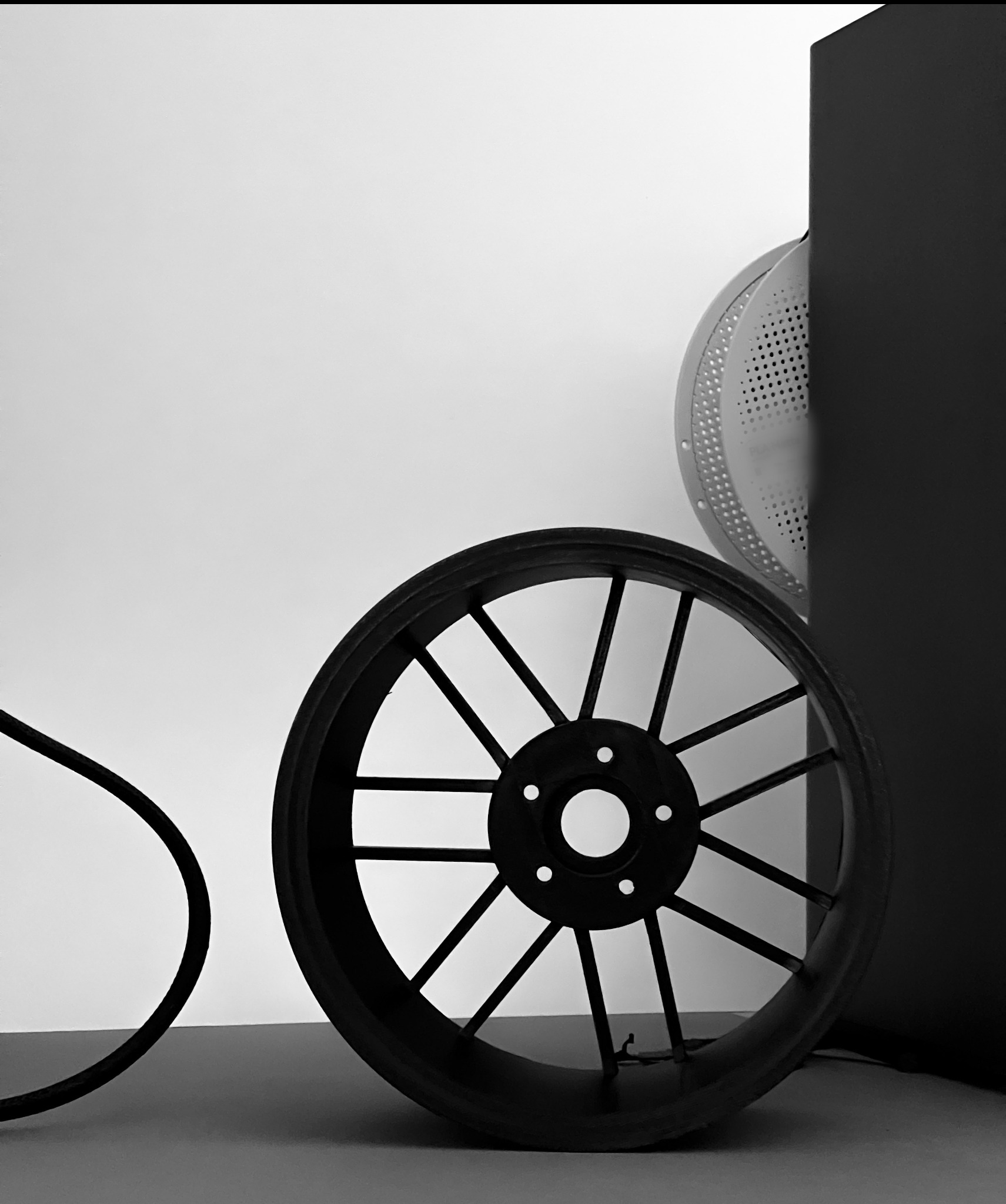 Czarne koło z tworzywa sztucznego z ażurową konstrukcją, obok fragment bębna pralki i prostopadłościanu. Monochromatyczne ujęcie detali.