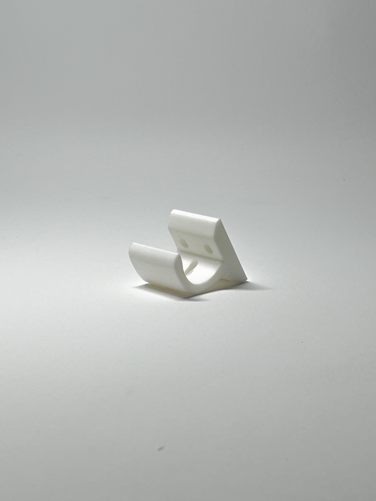 Biały, plastikowy uchwyt, wydrukowany w 3D, leży na białym tle. Widoczne otwory montażowe i delikatna tekstura druku warstwowego.