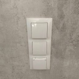 MONTE KABLO USŁUGI ELEKTRYCZNE DOMINIK KAPTUR - Biały, potrójny włącznik światła z gniazdkiem w szarej ścianie. Detal instalacji elektrycznej w domu w Czempiniu. Nowoczesny design.