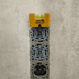 MONTE KABLO USŁUGI ELEKTRYCZNE DOMINIK KAPTUR - Gniazdka elektryczne w szarej ścianie, poziomica Stabila Pocket Electric na górze, widoczne elementy montażowe, instalacja w trakcie, neutralne oświetlenie.