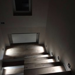 MONTE KABLO USŁUGI ELEKTRYCZNE DOMINIK KAPTUR - Schody z drewnianymi stopniami oświetlone punktowymi lampami ściennymi. Widok z góry na dół, okno dachowe w tle.