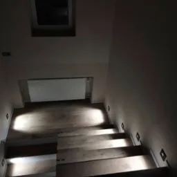 Schody z drewnianymi stopniami oświetlone punktowymi lampami ściennymi. Widok z góry na dół, okno dachowe w tle.