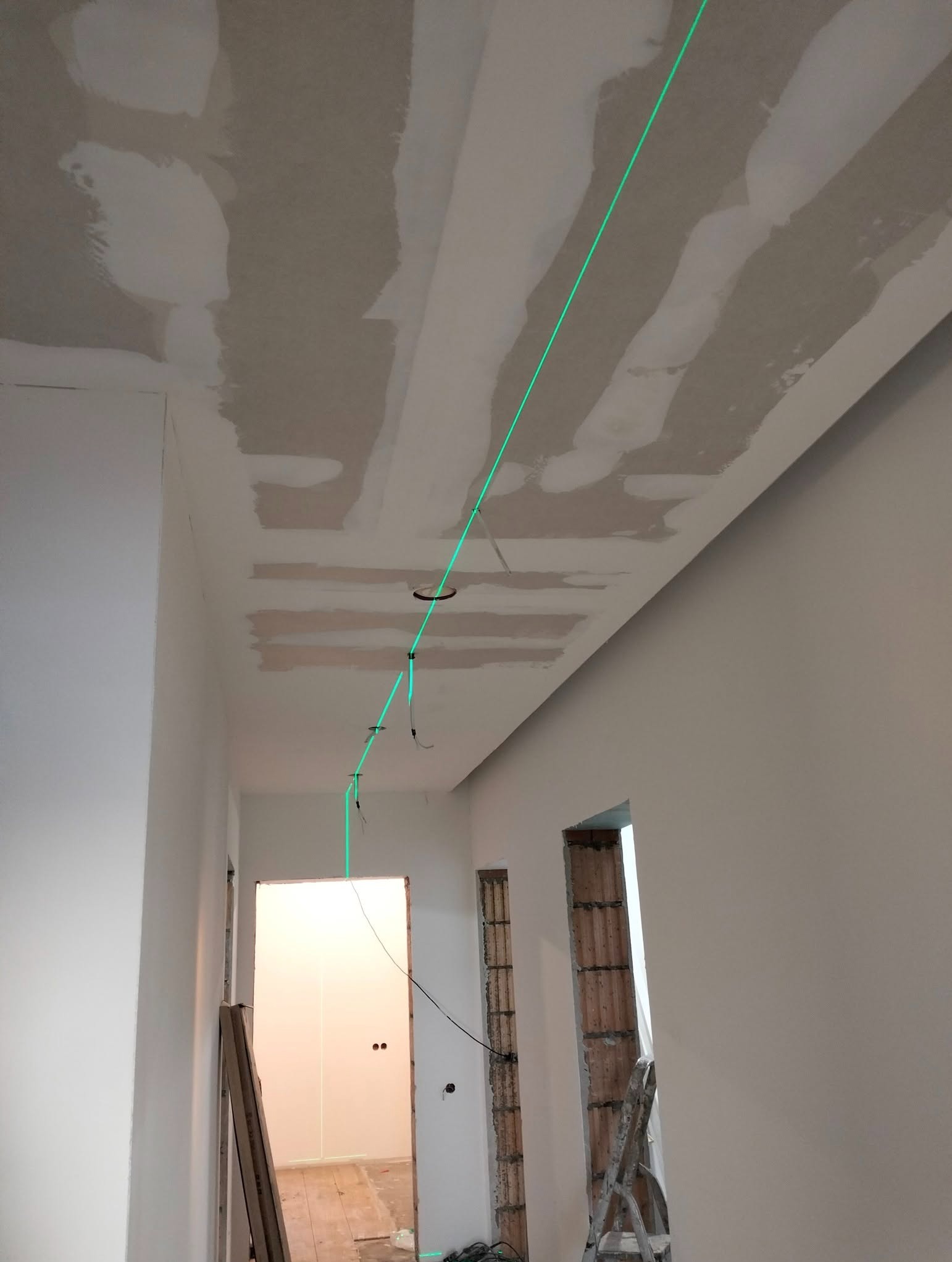 Zielona linia lasera wyznacza punkty montażu oświetlenia na suficie z płyt gipsowo-kartonowych w korytarzu. Widoczne przewody i otwory pod lampy.