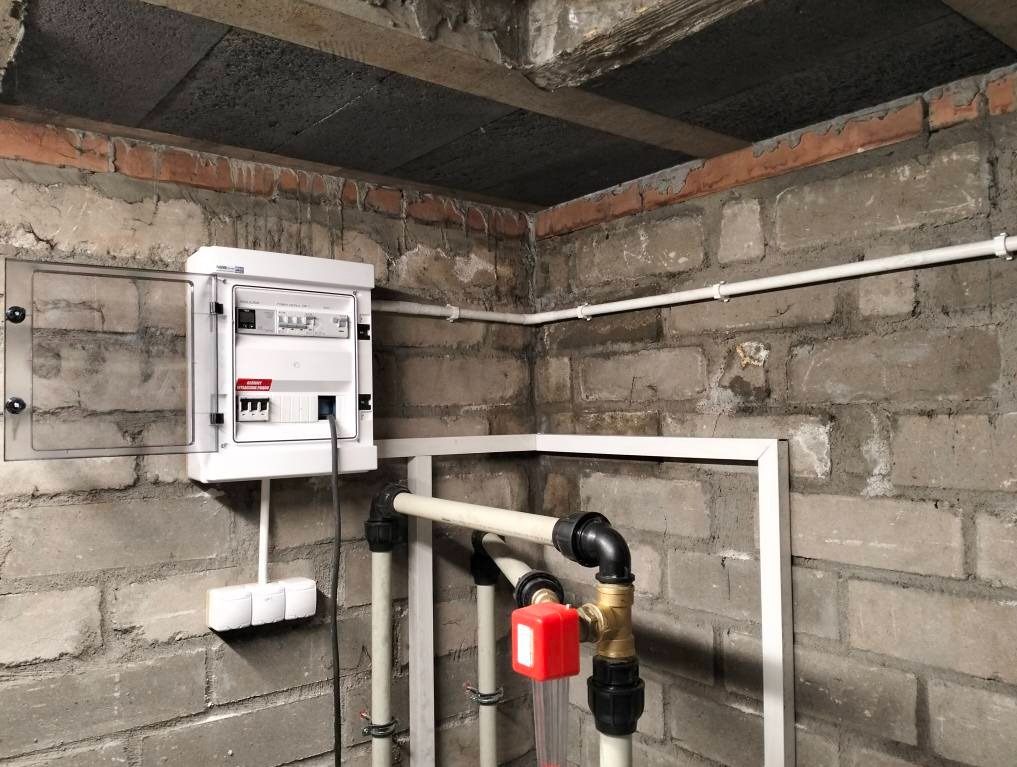 Rozdzielnica elektryczna z otwartymi drzwiczkami na tle szarej ściany z cegły. Widoczne rury instalacyjne i przewody. Ujęcie w piwnicy lub pomieszczeniu gospodarczym.