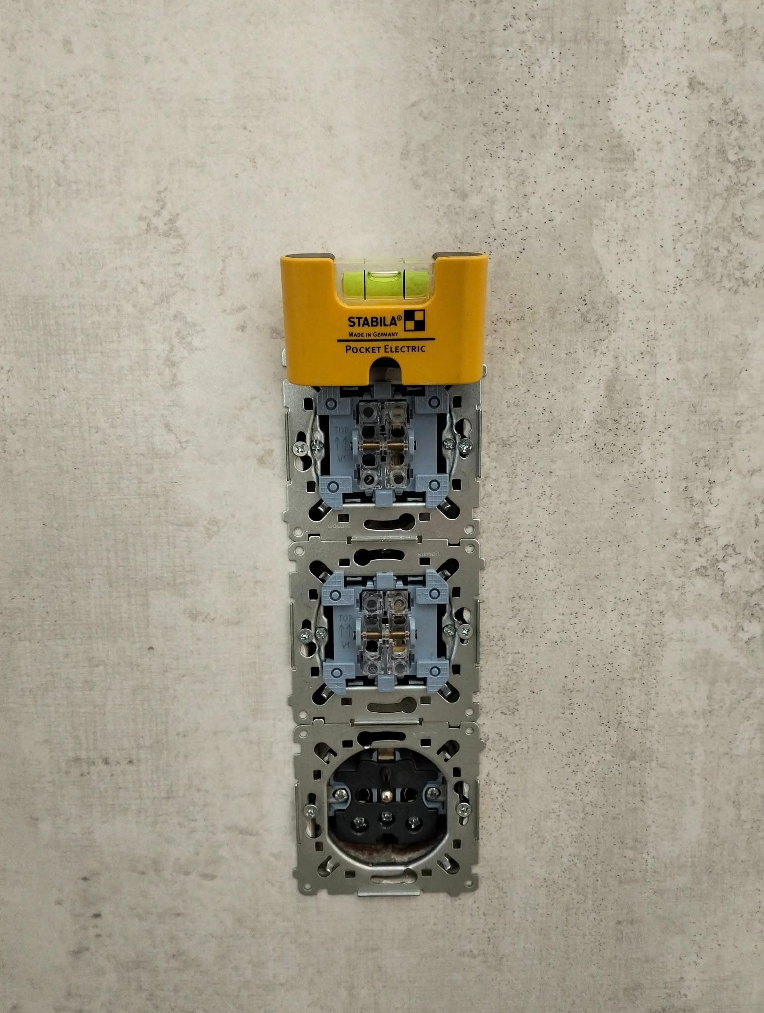 Gniazdka elektryczne w szarej ścianie, poziomica Stabila Pocket Electric na górze, widoczne elementy montażowe, instalacja w trakcie, neutralne oświetlenie.
