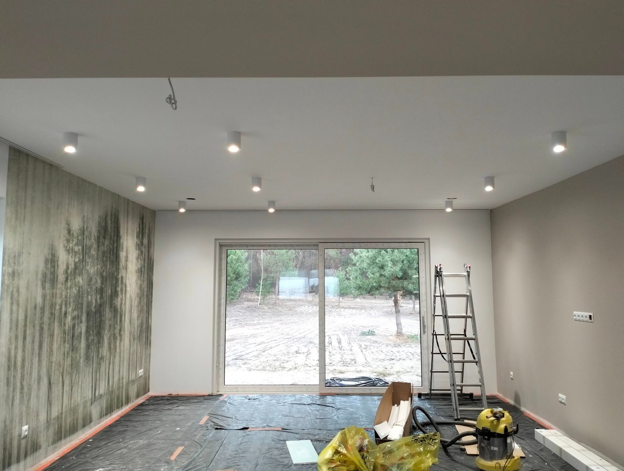 Wnętrze pokoju z zamontowanym oświetleniem sufitowym typu spot. Widoczne przewody do podłączenia lamp, drabina, okno tarasowe i podłoga zabezpieczona folią.