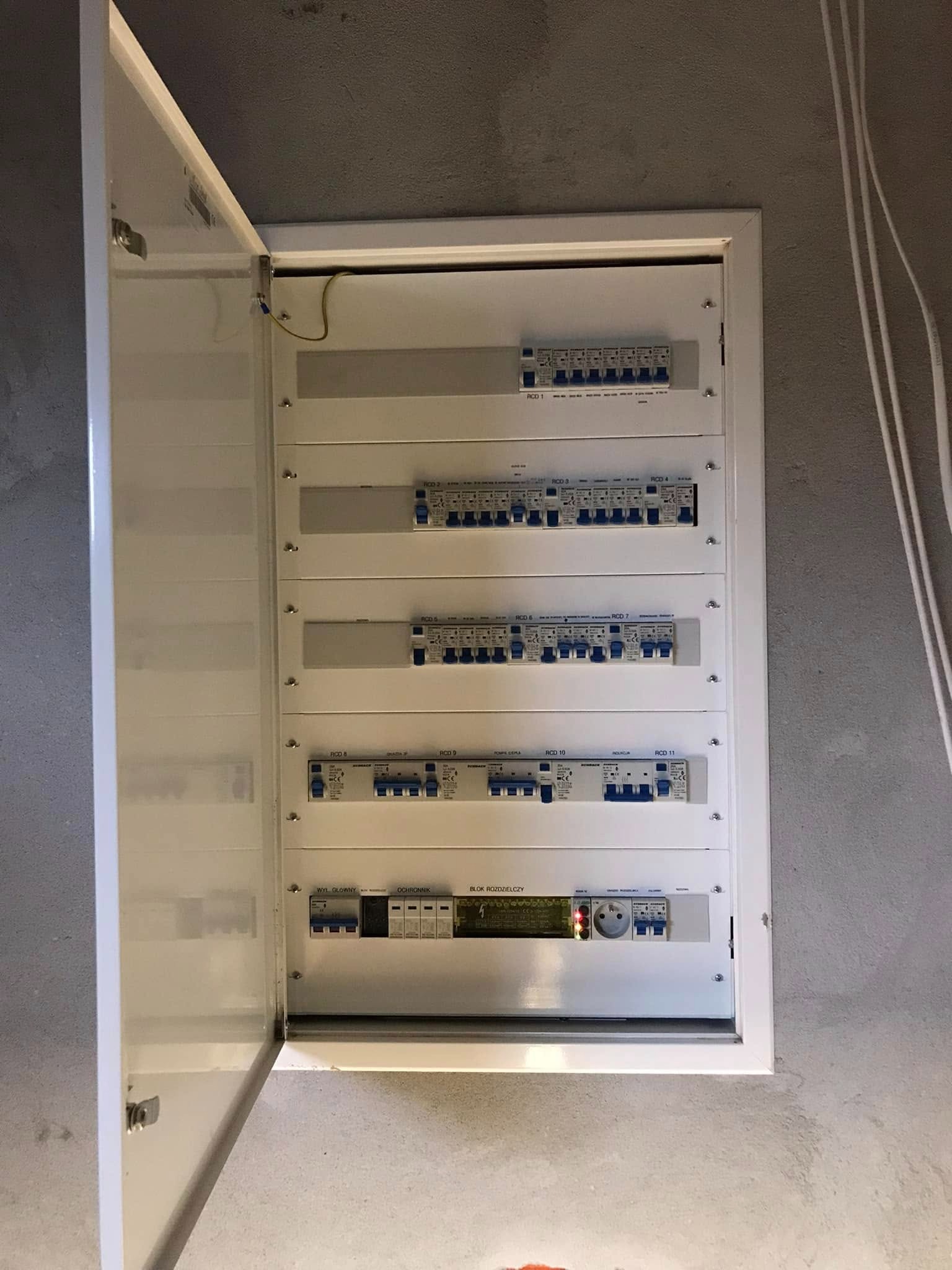 Otwarta, biała skrzynka rozdzielcza z bezpiecznikami na szarej, surowej ścianie. Widoczne przewody elektryczne. Dolny moduł z gniazdem i diodami.