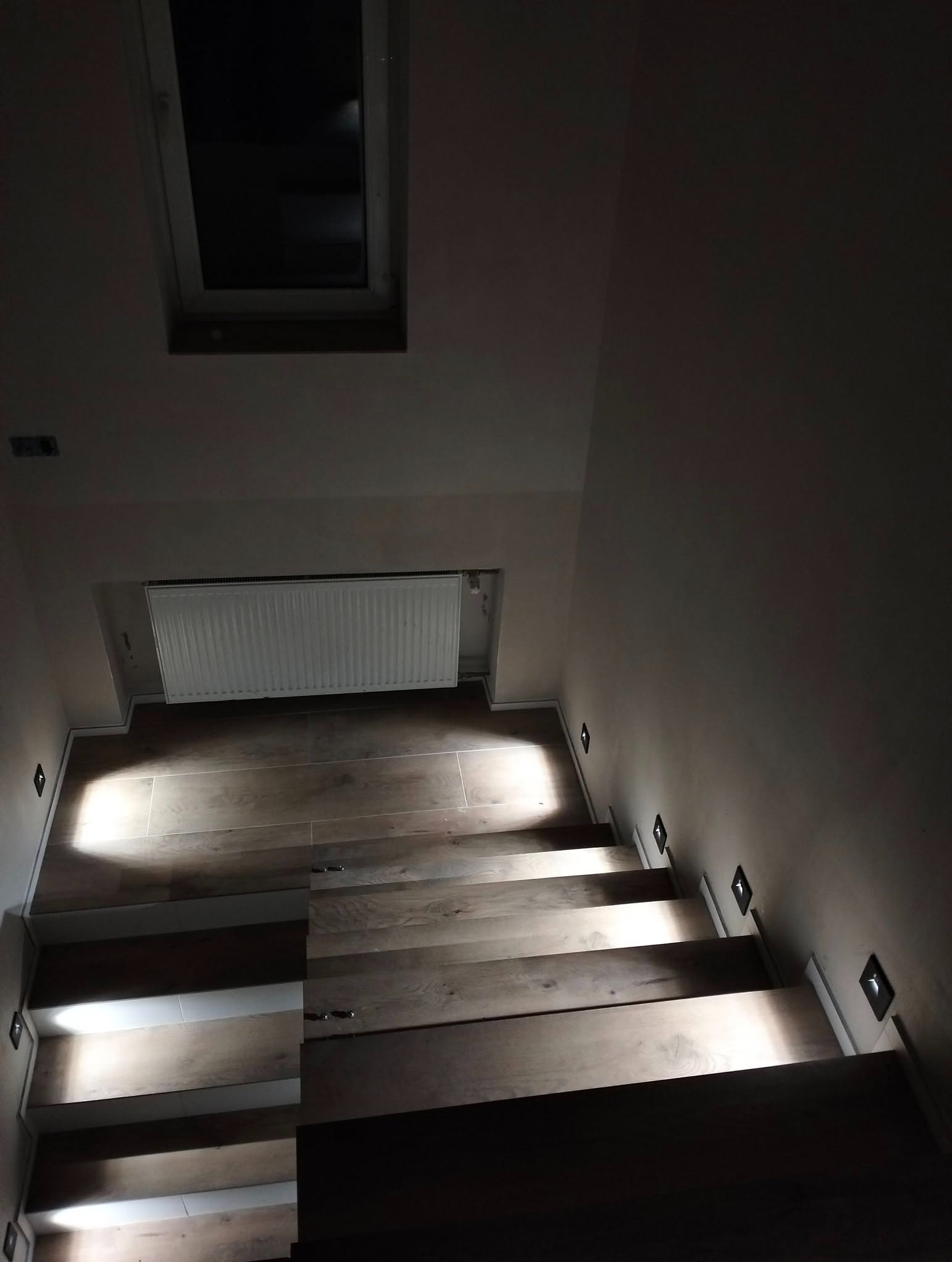 Schody z drewnianymi stopniami oświetlone punktowymi lampami ściennymi. Widok z góry na dół, okno dachowe w tle.