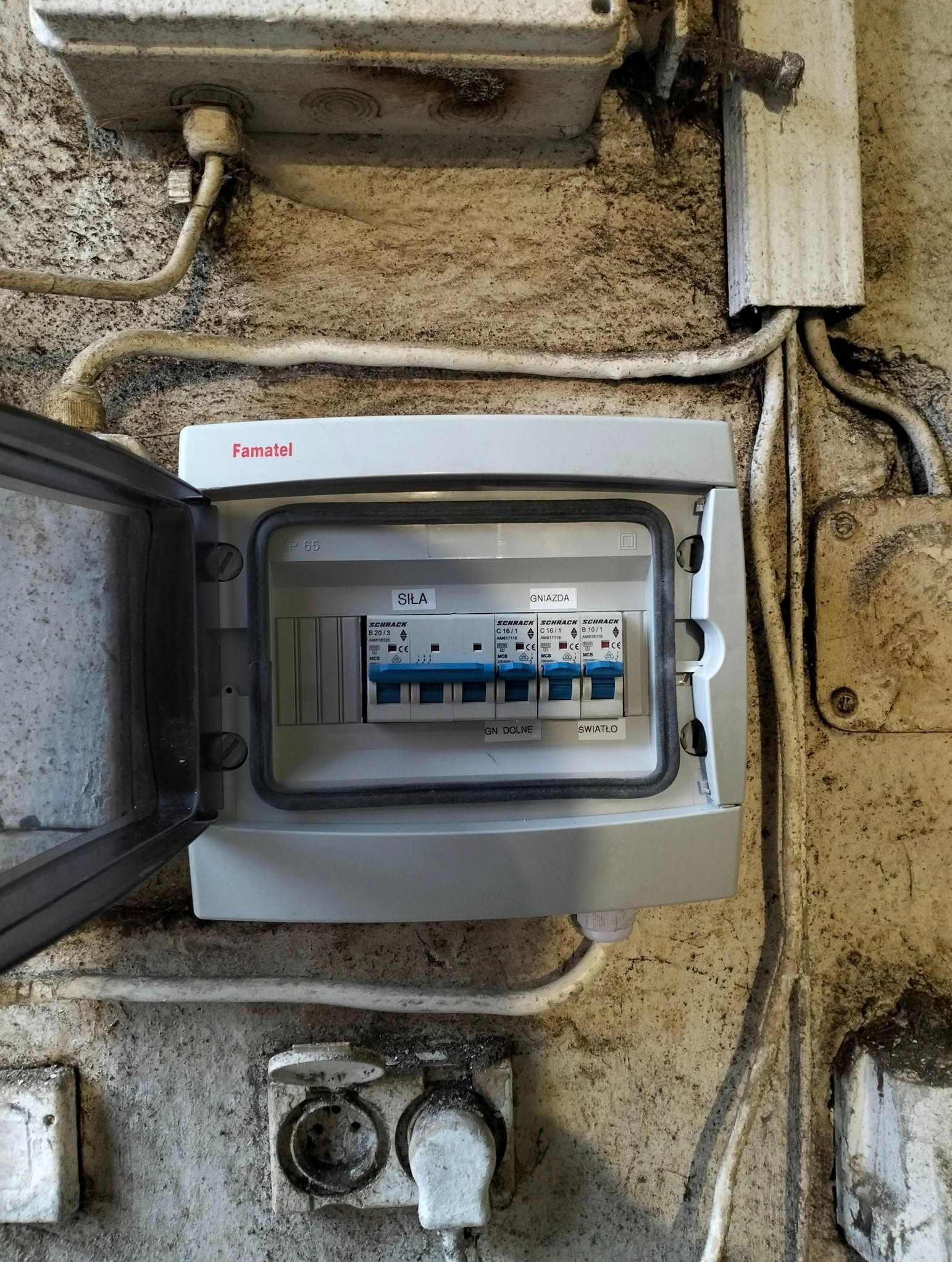 Otwarta szara skrzynka elektryczna marki Famatel z wyłącznikami Schrack na brudnej ścianie. Widoczne przewody i gniazda elektryczne.
