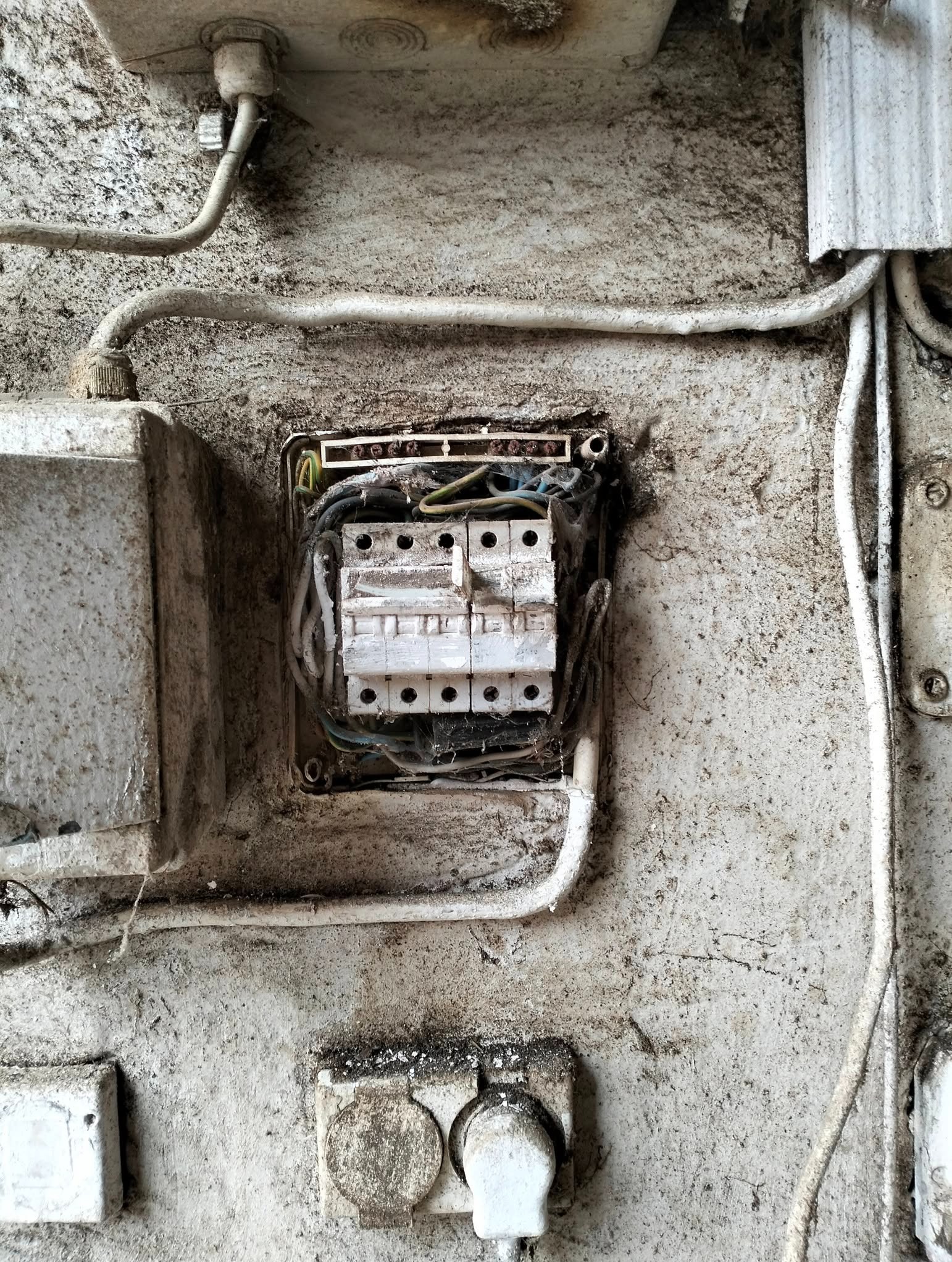 Stary, zaniedbany rozdzielacz elektryczny z przewodami na brudnej, pomalowanej ścianie. Widoczne bezpieczniki i gniazdka. Wnętrze rozdzielacza zakurzone i zaśniedziałe.