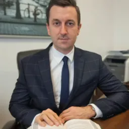 Portret adwokata w biurze, siedzącego za biurkiem z otwartą książką, w tle obraz z motywem miejskim i drukarka. Mężczyzna w garniturze patrzy wprost.