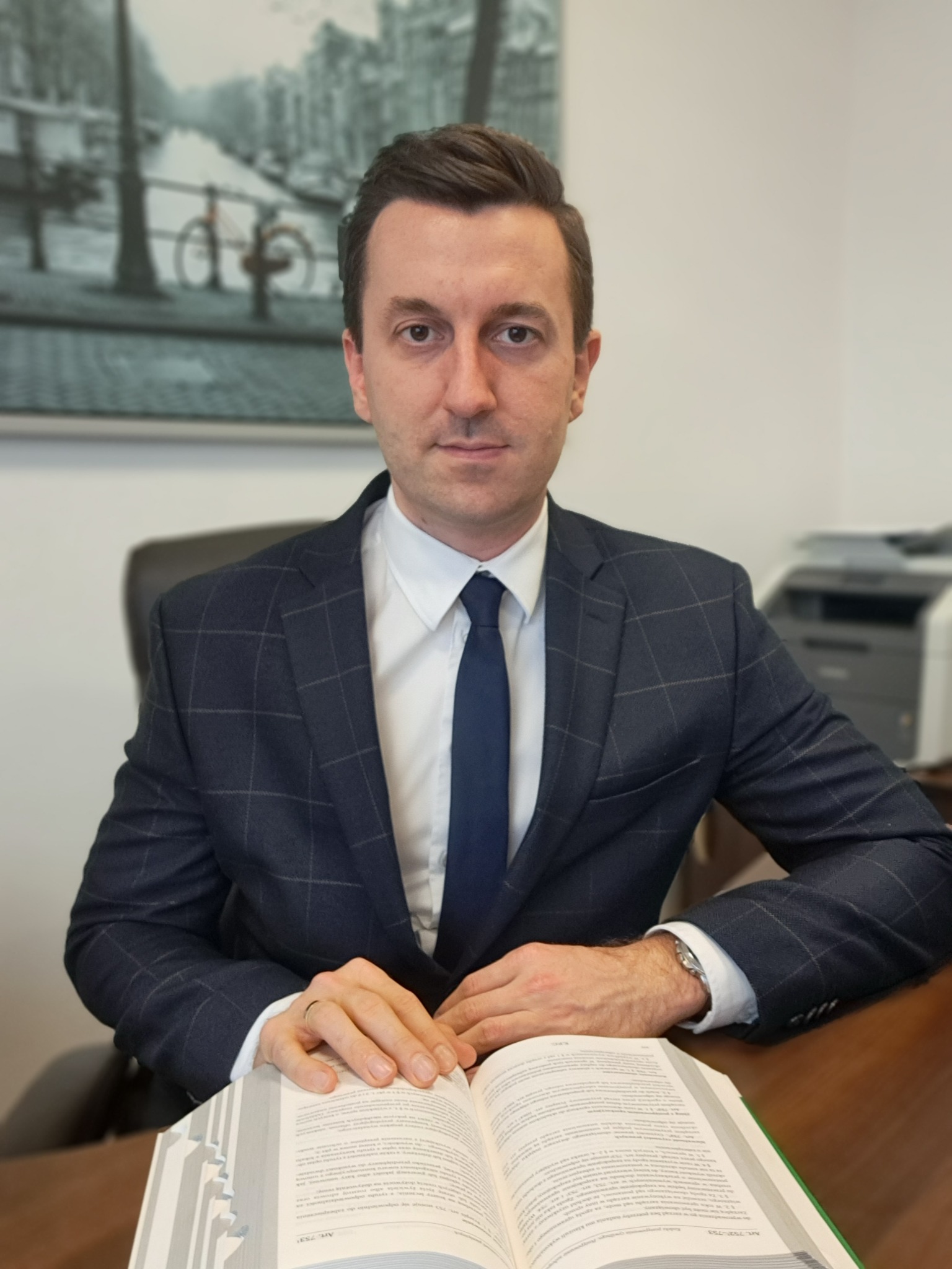 Portret adwokata w biurze, siedzącego za biurkiem z otwartą książką, w tle obraz z motywem miejskim i drukarka. Mężczyzna w garniturze patrzy wprost.