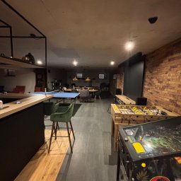 MARCIN GONTARCZYK USŁUGI OGÓLNOBUDOWLANE - Kącik rozrywki w piwnicy: stół do ping-ponga, piłkarzyki i fliper Jurassic Park. Ceglana ściana, bar z hokerami, oświetlenie punktowe. Nowoczesny design.