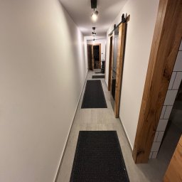 MARCIN GONTARCZYK USŁUGI OGÓLNOBUDOWLANE - Długi korytarz z białymi ścianami, szarymi płytkami podłogowymi i czarnymi dywanikami. Widoczne drewniane ramy drzwi przesuwnych i nowoczesne oświetlenie sufitowe.