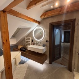 MARCIN GONTARCZYK USŁUGI OGÓLNOBUDOWLANE - Jasna łazienka na poddaszu z drewnianymi belkami, okrągłym lustrem z podświetleniem LED i marmurową podłogą. Widok na otwarte drzwi do dalszej części domu.