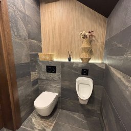 MARCIN GONTARCZYK USŁUGI OGÓLNOBUDOWLANE - Nowoczesna łazienka z szarymi płytkami imitującymi kamień, podświetlaną ścianą z drewnianymi lamelami i wiszącą toaletą oraz pisuar. Minimalistyczny design.