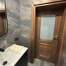 MARCIN GONTARCZYK USŁUGI OGÓLNOBUDOWLANE - Nowoczesna łazienka z szarymi płytkami imitującymi kamień, drewnianymi drzwiami z matową szybą i czarną szafką pod umywalką z blatem. Minimalistyczny design.