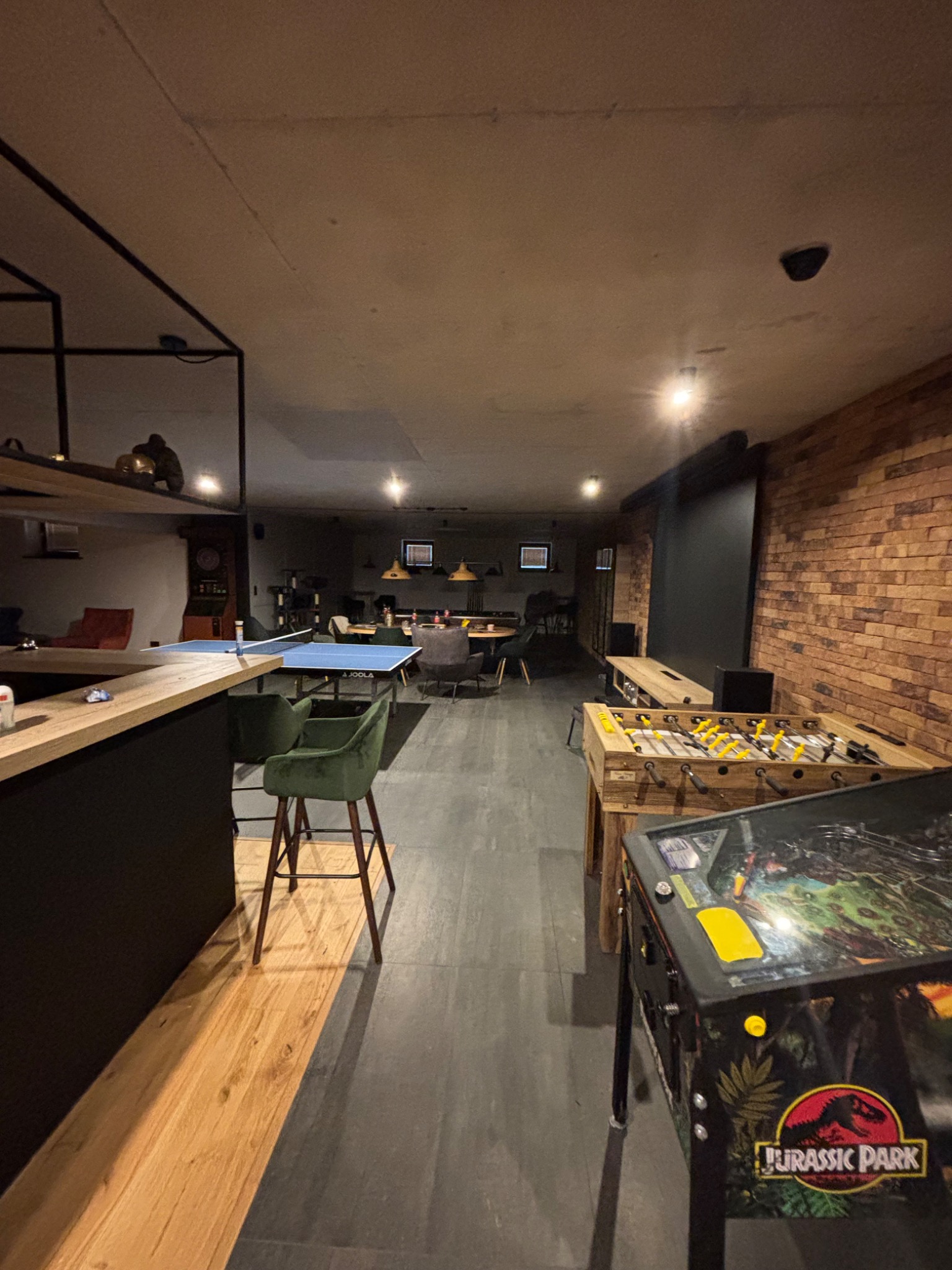 Kącik rozrywki w piwnicy: stół do ping-ponga, piłkarzyki i fliper Jurassic Park. Ceglana ściana, bar z hokerami, oświetlenie punktowe. Nowoczesny design.