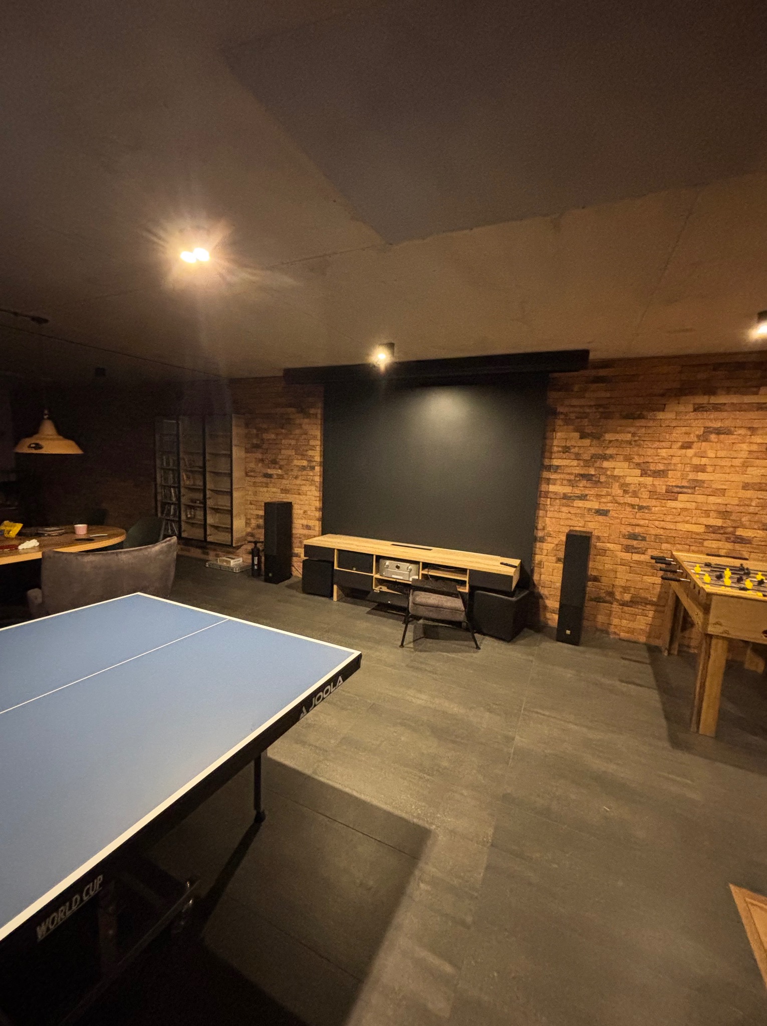Nowoczesna sala rozrywki z ceglaną ścianą, stołem do ping-ponga i piłkarzykami. Minimalistyczny design, ciemna podłoga, oświetlenie punktowe. Widoczny ekran projekcyjny.