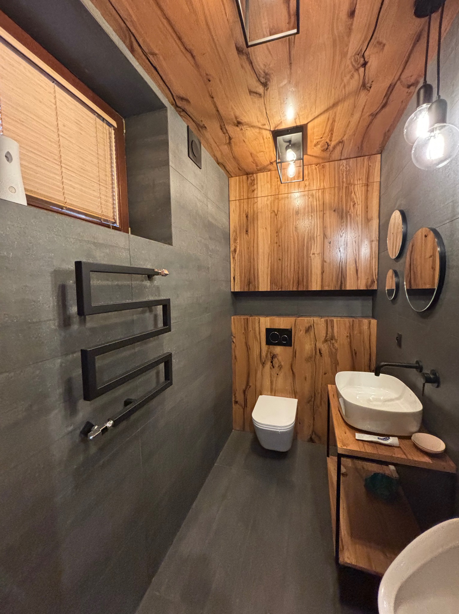 Nowoczesna łazienka z drewnianym sufitem i elementami wykończenia. Szare płytki, podwieszana toaleta i designerski grzejnik drabinkowy.