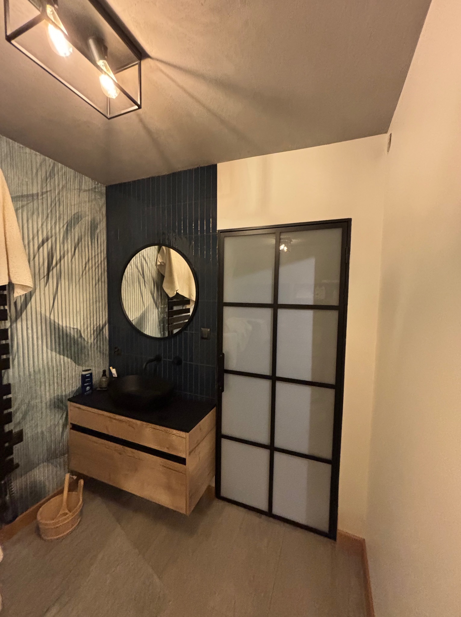 Nowoczesna łazienka z umywalką nablatową, okrągłym lustrem i kabiną prysznicową w stylu loft. Ciemne płytki i drewniane elementy dodają elegancji.