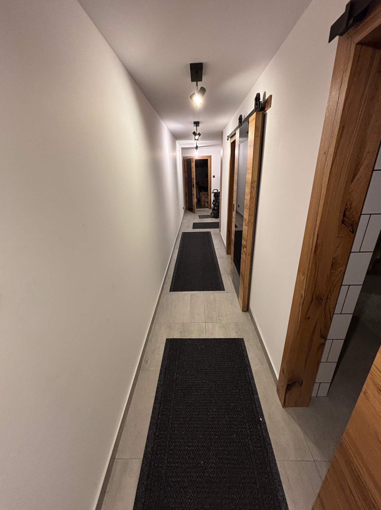 Długi korytarz z białymi ścianami, szarymi płytkami podłogowymi i czarnymi dywanikami. Widoczne drewniane ramy drzwi przesuwnych i nowoczesne oświetlenie sufitowe.