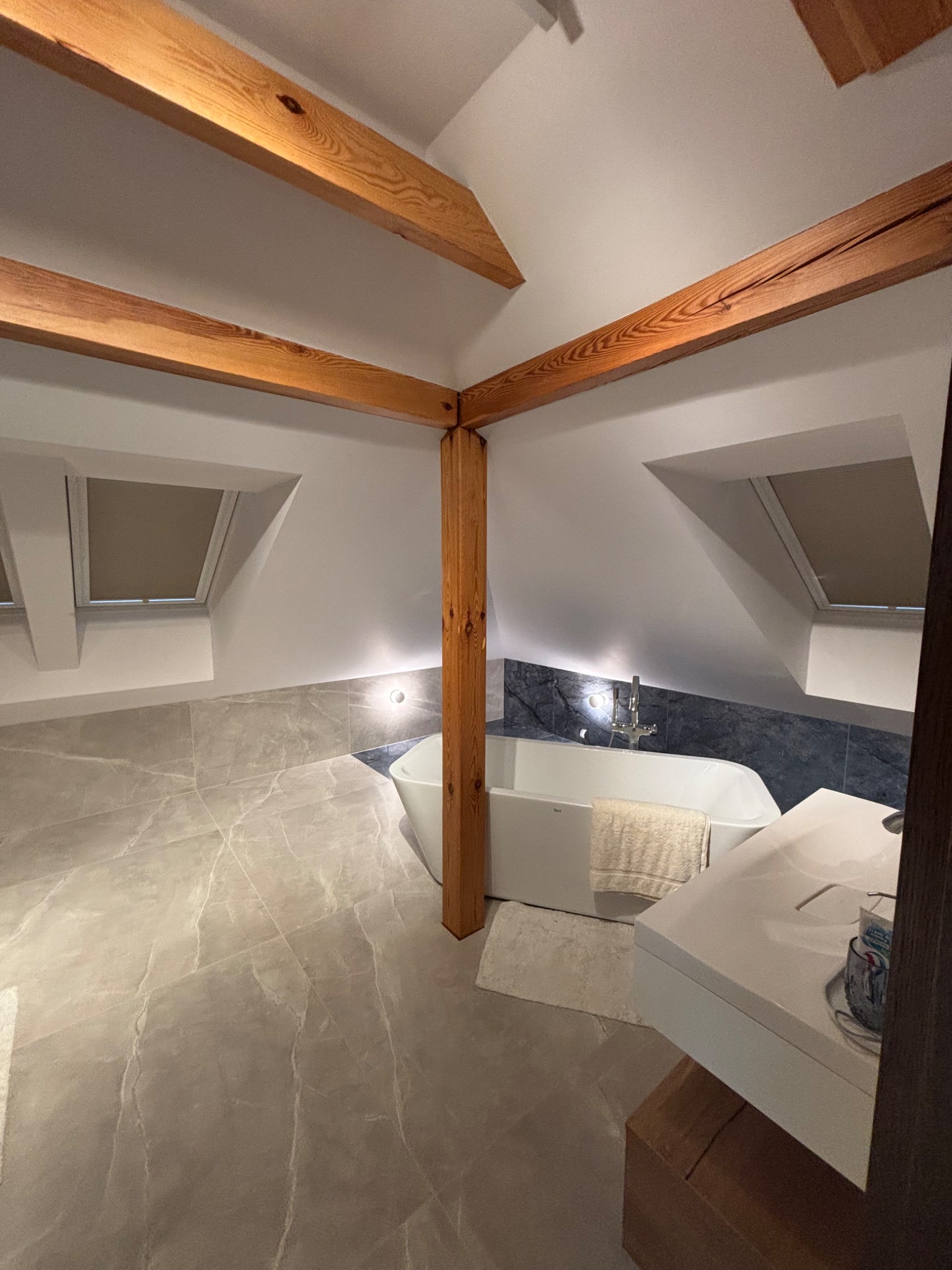 Jasna łazienka na poddaszu z drewnianymi belkami stropowymi, szarą marmurową podłogą i ścianą za wanną. Nowoczesna armatura i okna dachowe.