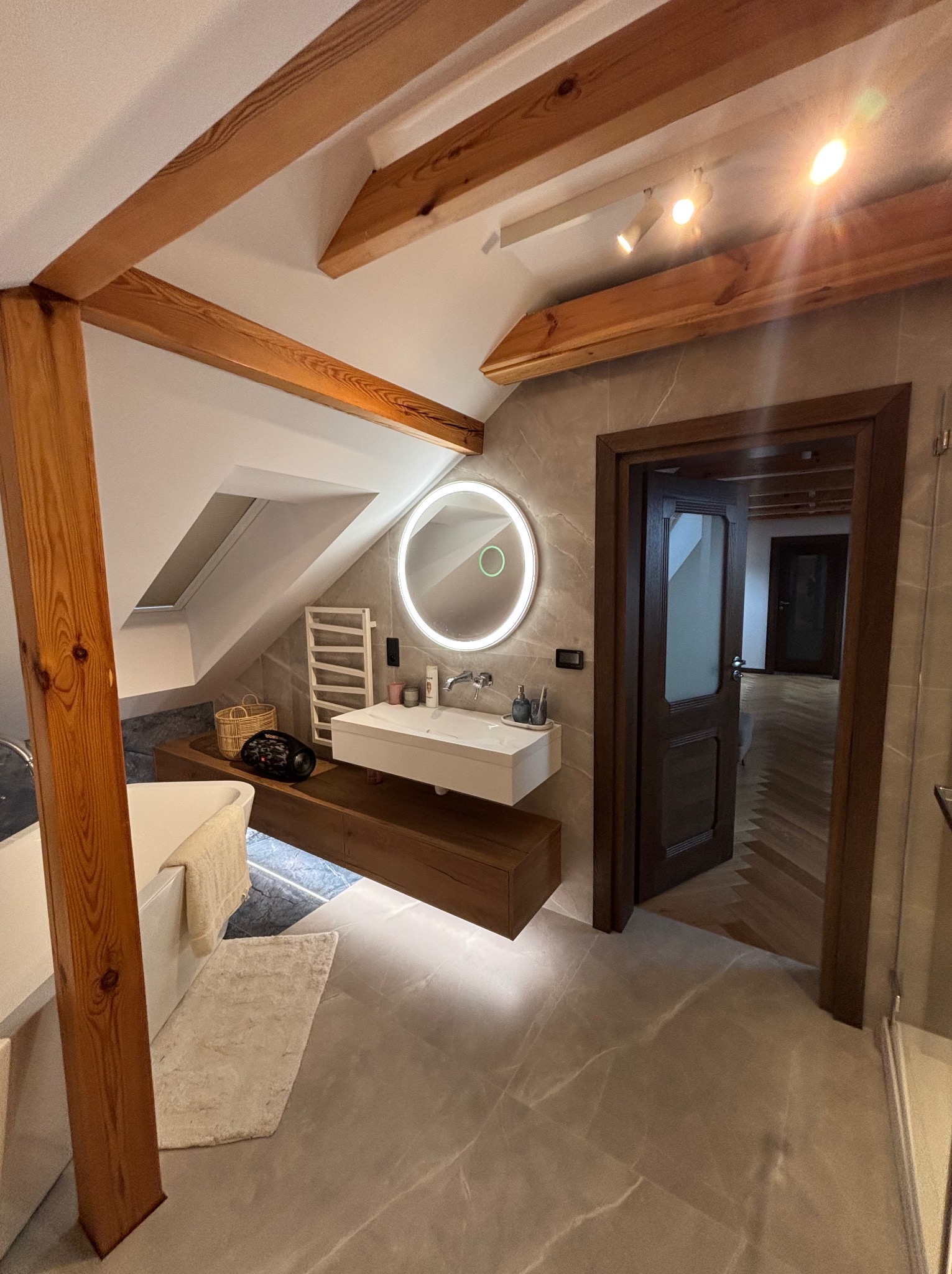 Jasna łazienka na poddaszu z drewnianymi belkami, okrągłym lustrem z podświetleniem LED i marmurową podłogą. Widok na otwarte drzwi do dalszej części domu.
