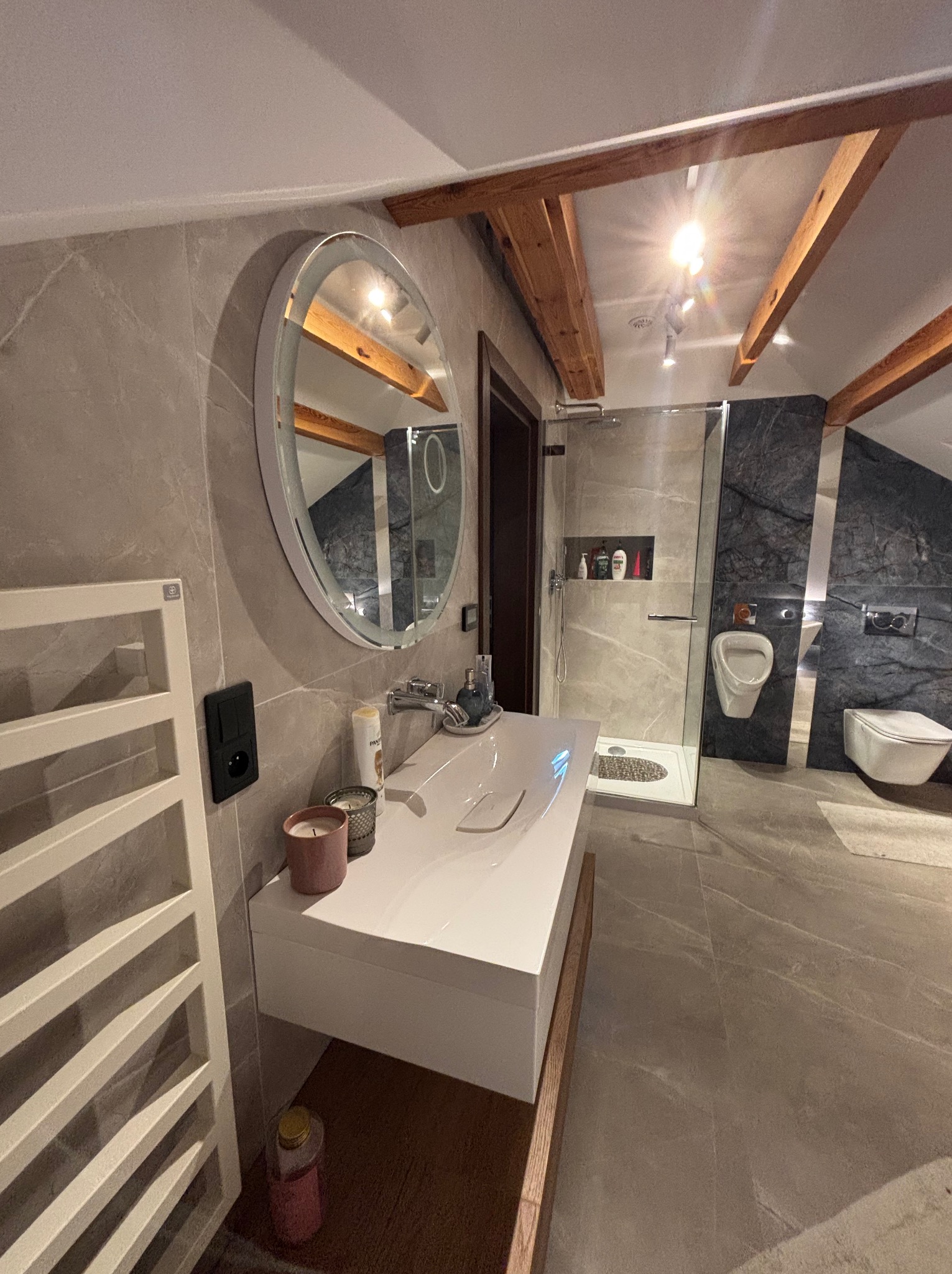 Nowoczesna łazienka z szarą marmurową płytką, drewnianymi belkami stropowymi i okrągłym lustrem z podświetleniem. Widoczna kabina prysznicowa i toaleta.