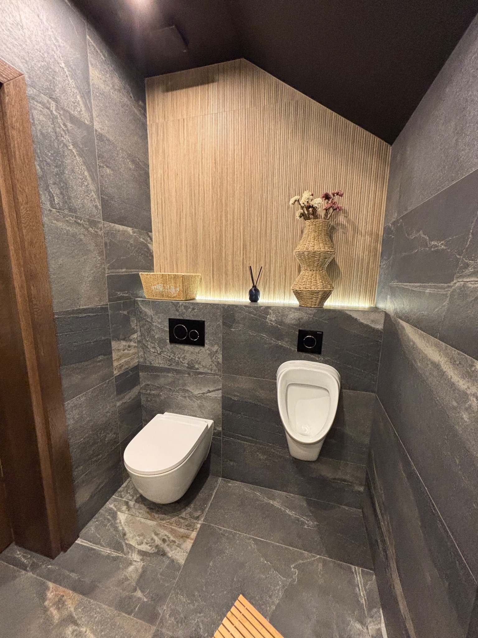 Nowoczesna łazienka z szarymi płytkami imitującymi kamień, podświetlaną ścianą z drewnianymi lamelami i wiszącą toaletą oraz pisuar. Minimalistyczny design.