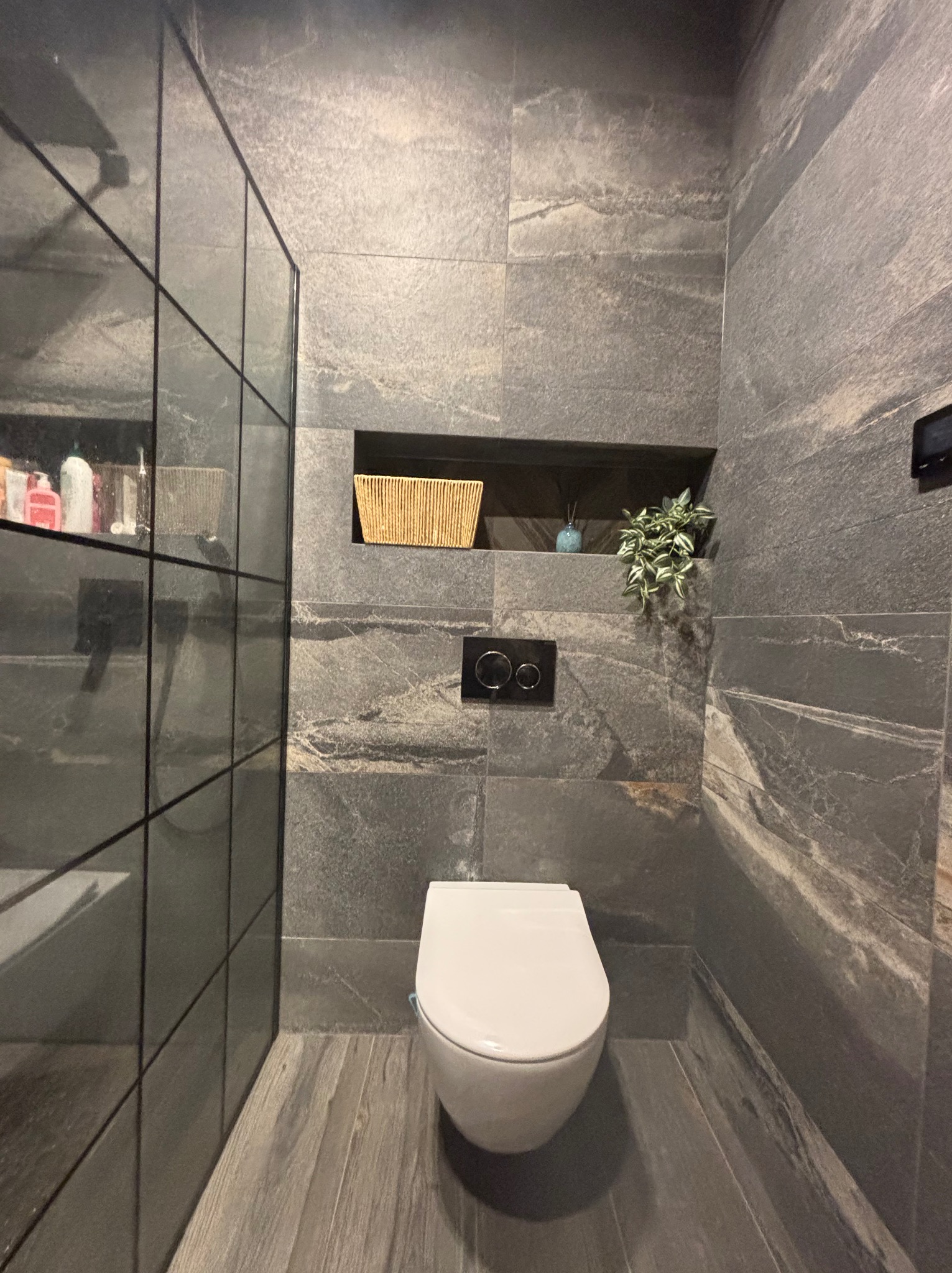 Nowoczesna łazienka z szarymi płytkami imitującymi kamień, wisząca toaleta, szklana kabina prysznicowa i wnęka z dekoracjami. Minimalistyczny design.