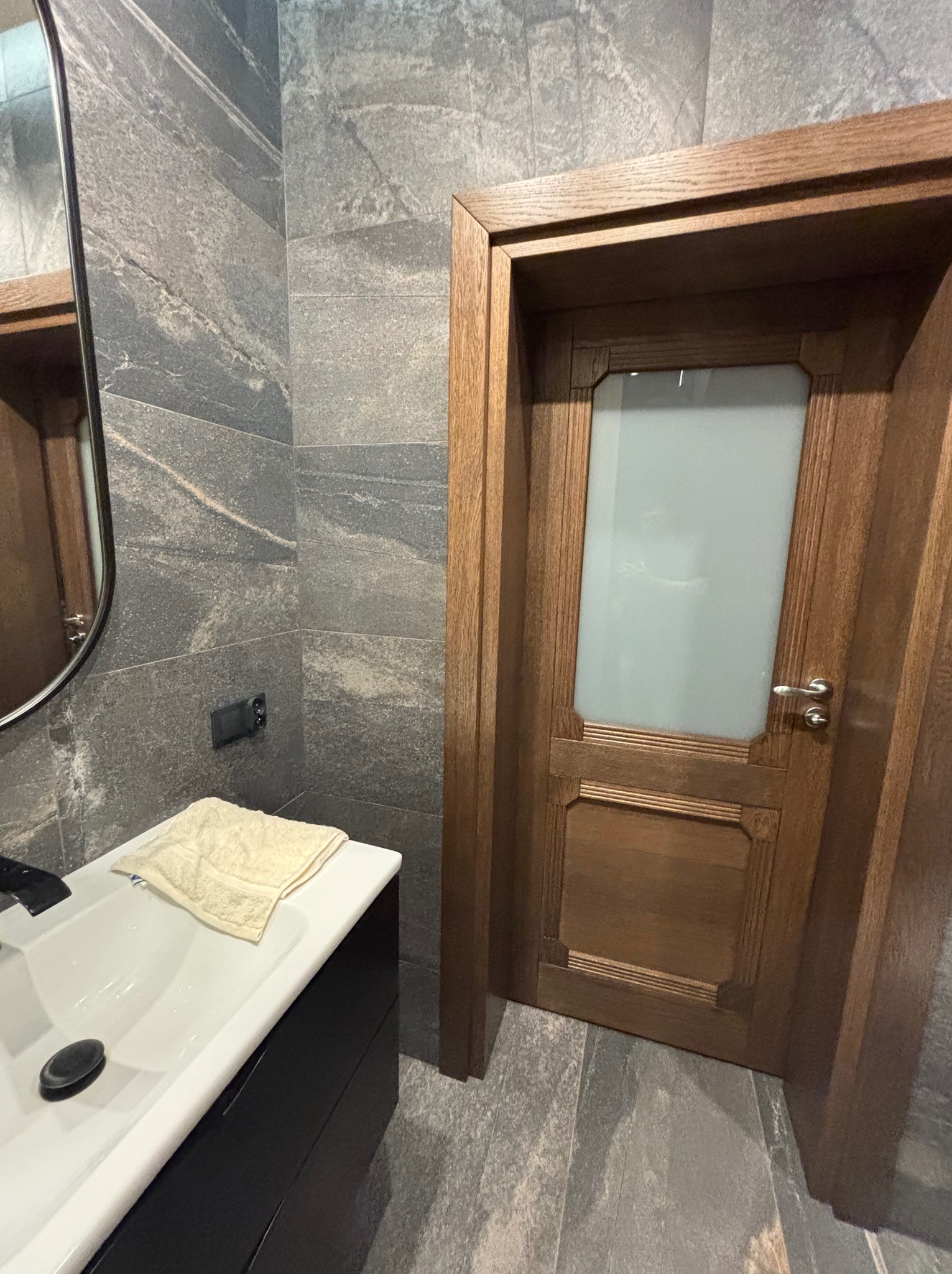 Nowoczesna łazienka z szarymi płytkami imitującymi kamień, drewnianymi drzwiami z matową szybą i czarną szafką pod umywalką z blatem. Minimalistyczny design.